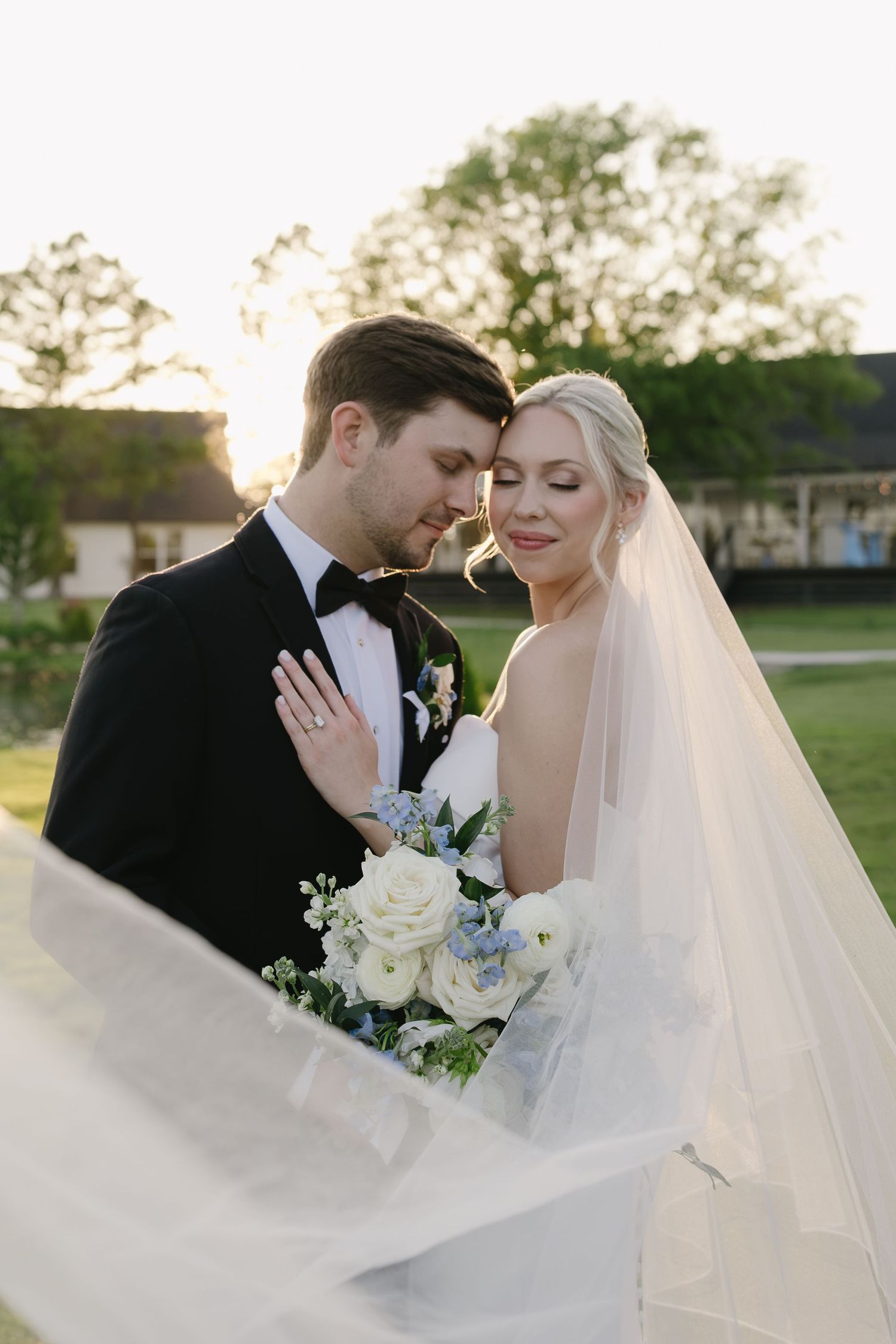 A bride and groom share a romantic golden hour moment, her veil flowing as she holds a white and blue bouquet.