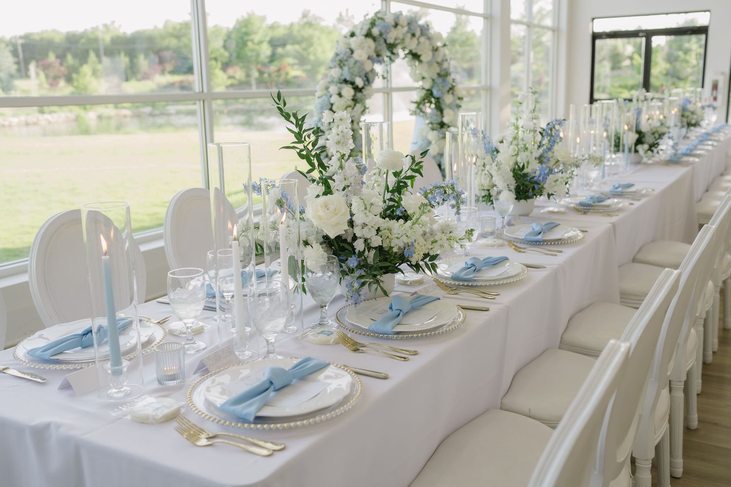 Elegant wedding reception table with white floral centerpieces, blue napkins, gold cutlery, and candles in a bright venue.