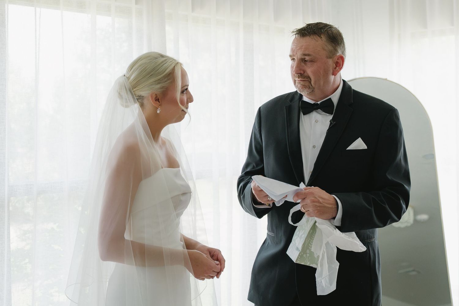 Bride in white gown and veil stands with groom in black tuxedo exchanging vows near a bright window.