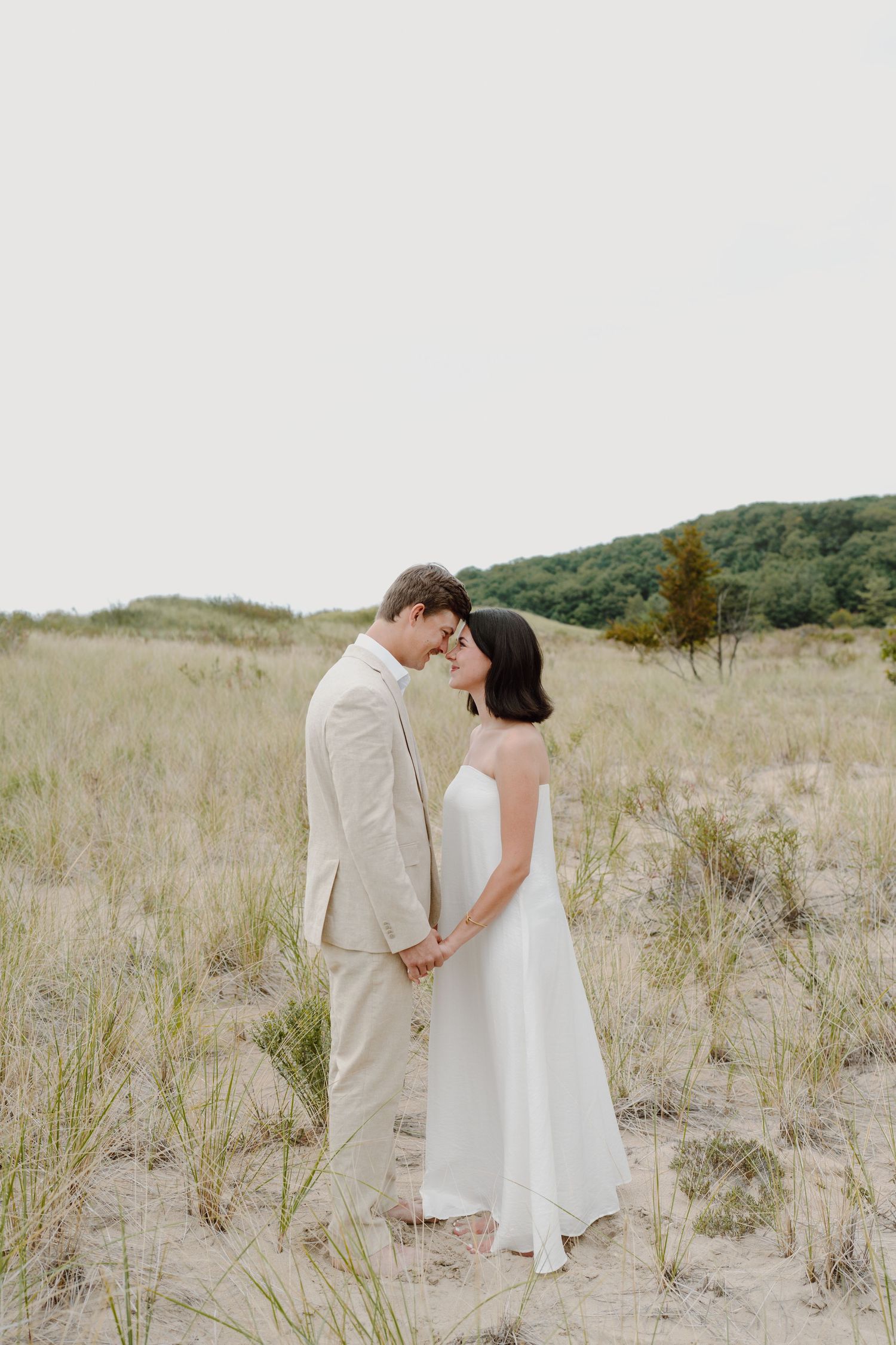 A tender moment between a couple in formal attire standing in a grassy dune landscape.