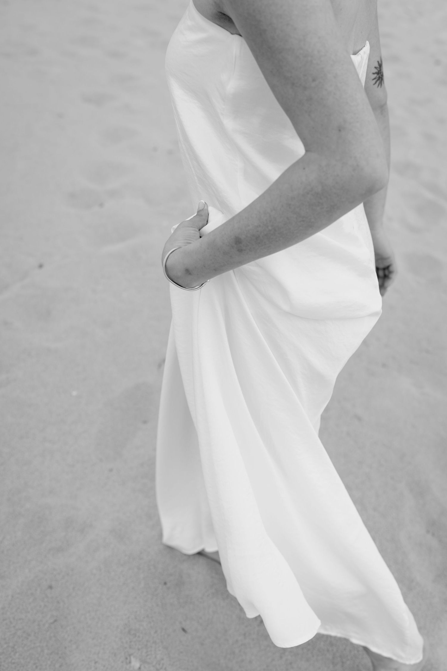 Close-up details of a flowing white wedding dress against sandy beach backdrop.