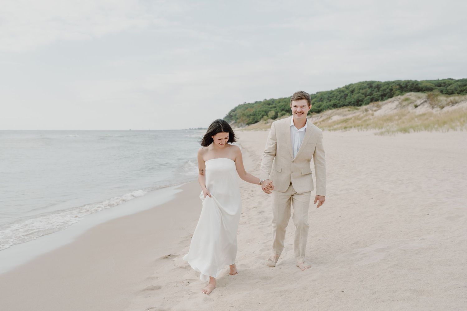 Elegant beach photo featuring a couple walking along the shoreline with dunes in the background.