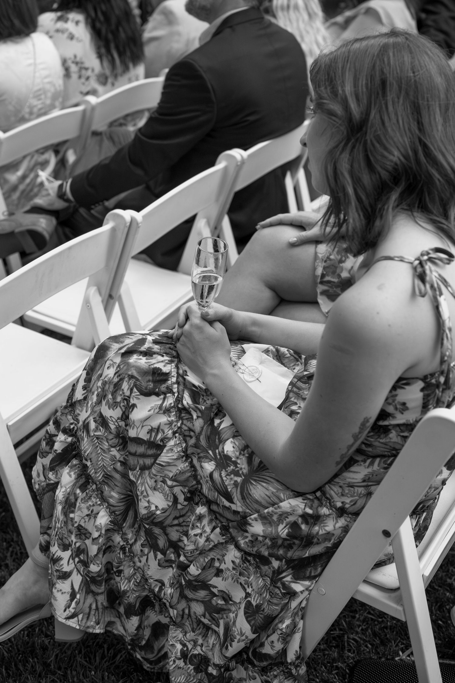 Close-up view of wedding guests seated in white folding chairs at an outdoor ceremony.