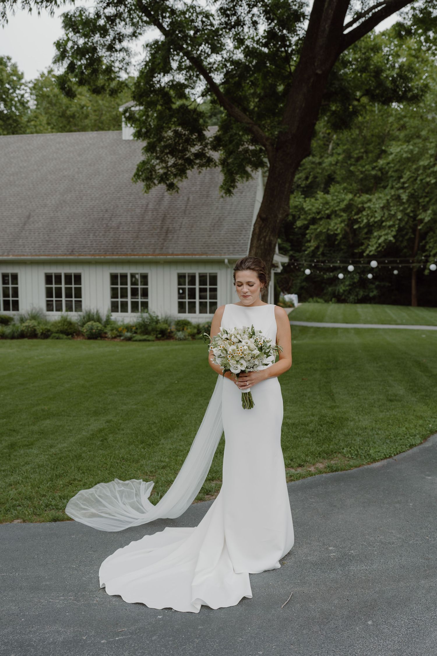 A bride in a flowing white dress with train poses on a paved path near a white building.