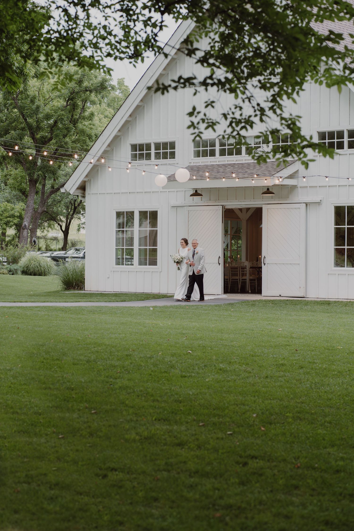Expansive lawn in front of a white barn wedding venue with string lights and trees.
