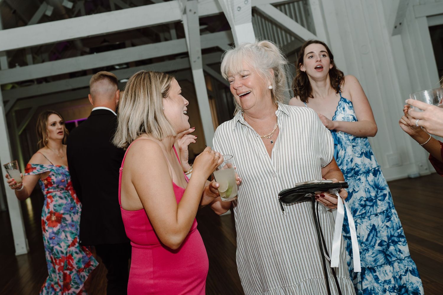 Wedding reception guests dance and socialize in a bright indoor venue with wooden beams.