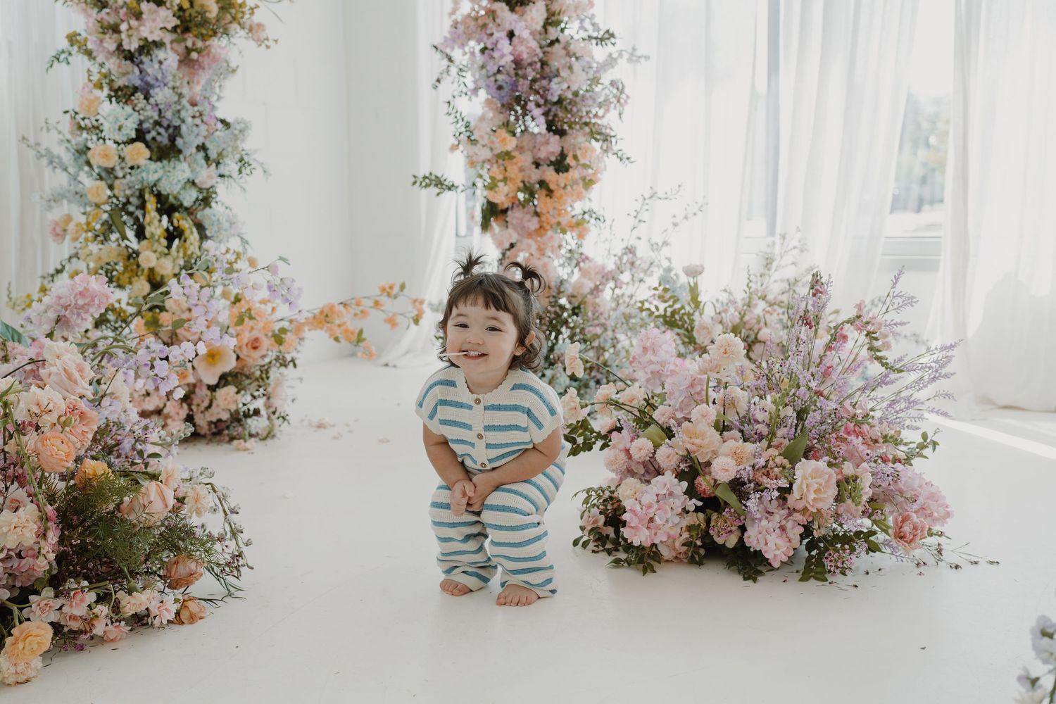 Toddler in blue and white striped outfit standing by pink and peach floral display.
