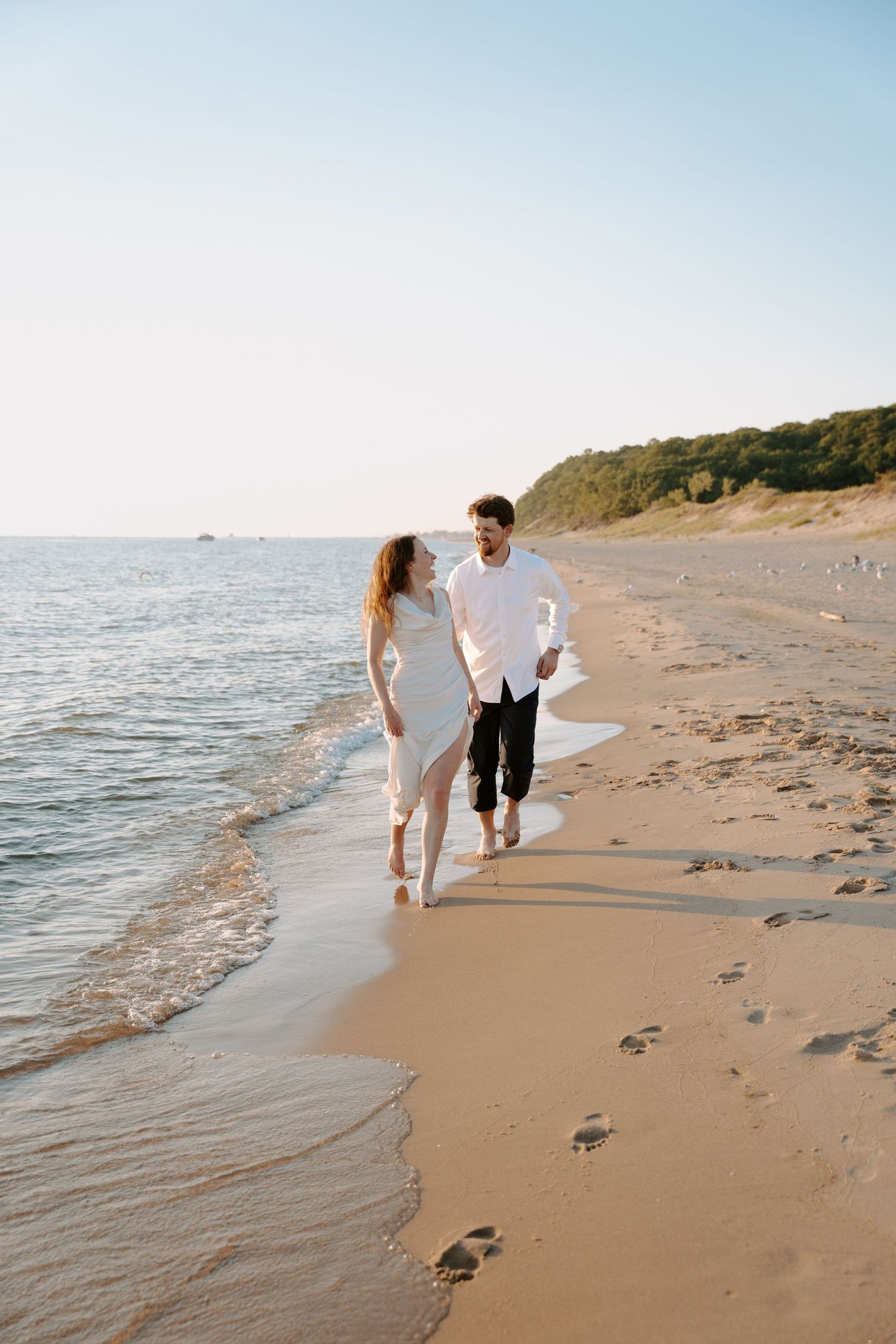 A couple strolls barefoot along a scenic beach shoreline during golden hour.