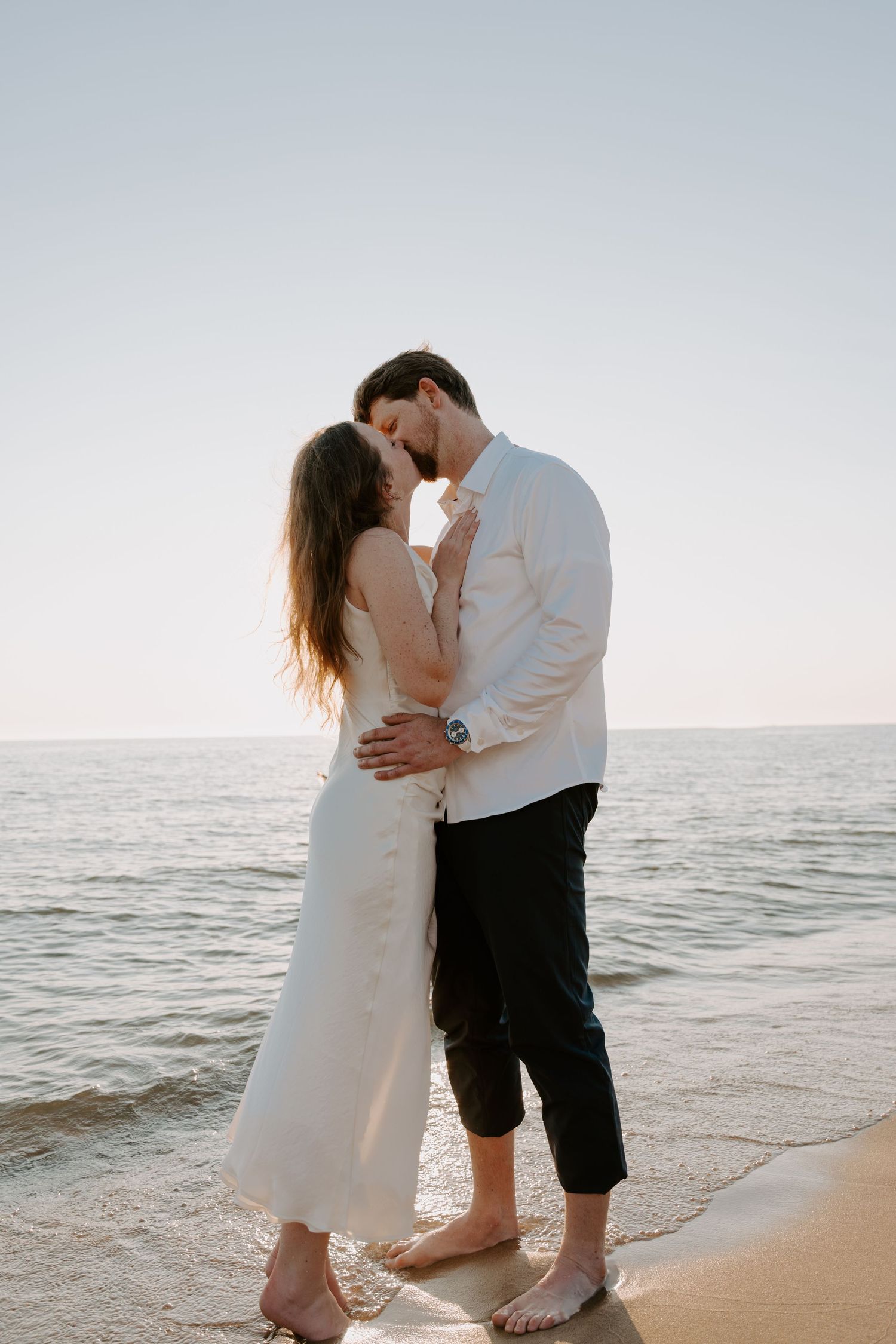 Tender embrace between couple on shoreline with calm ocean waters behind them.