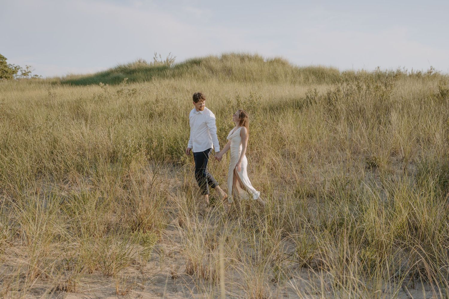 A couple explores a grassy coastal dune landscape under a bright sky.