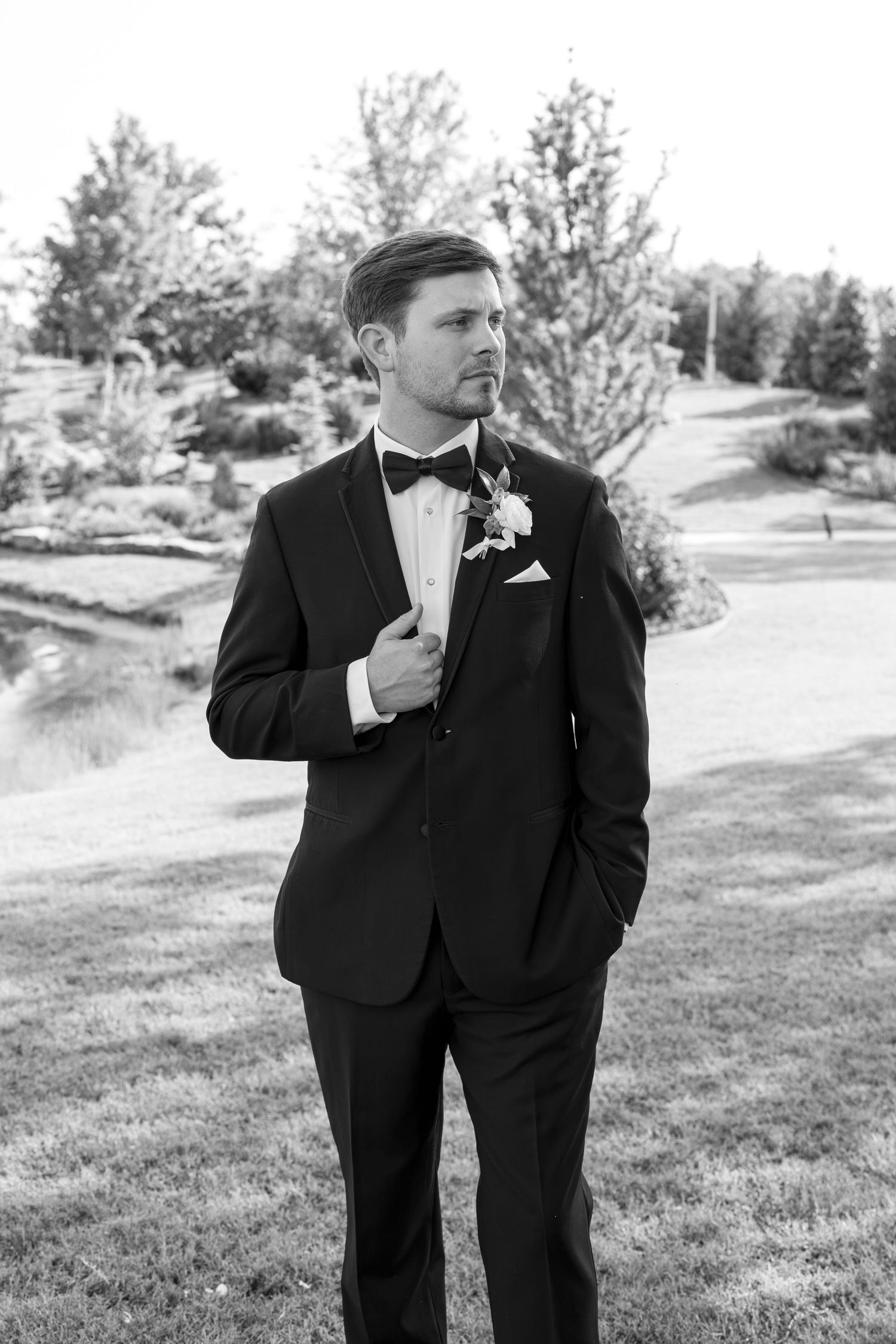 Groom in black tuxedo with bow tie and boutonniere poses outdoors near a pond in a black and white wedding photo.