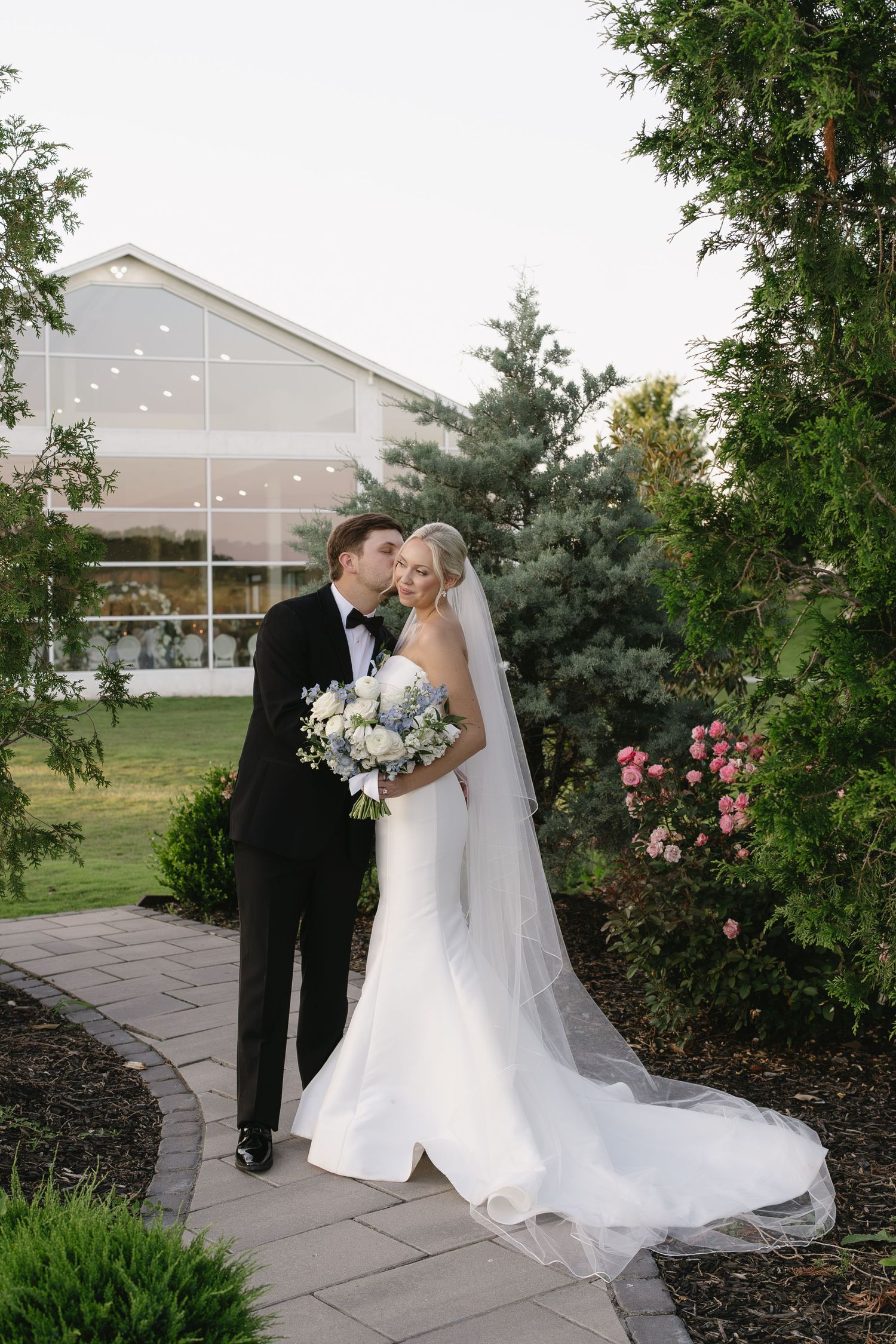 Bride and groom share a romantic moment on a garden path, with a white wedding venue and lush greenery in the background.