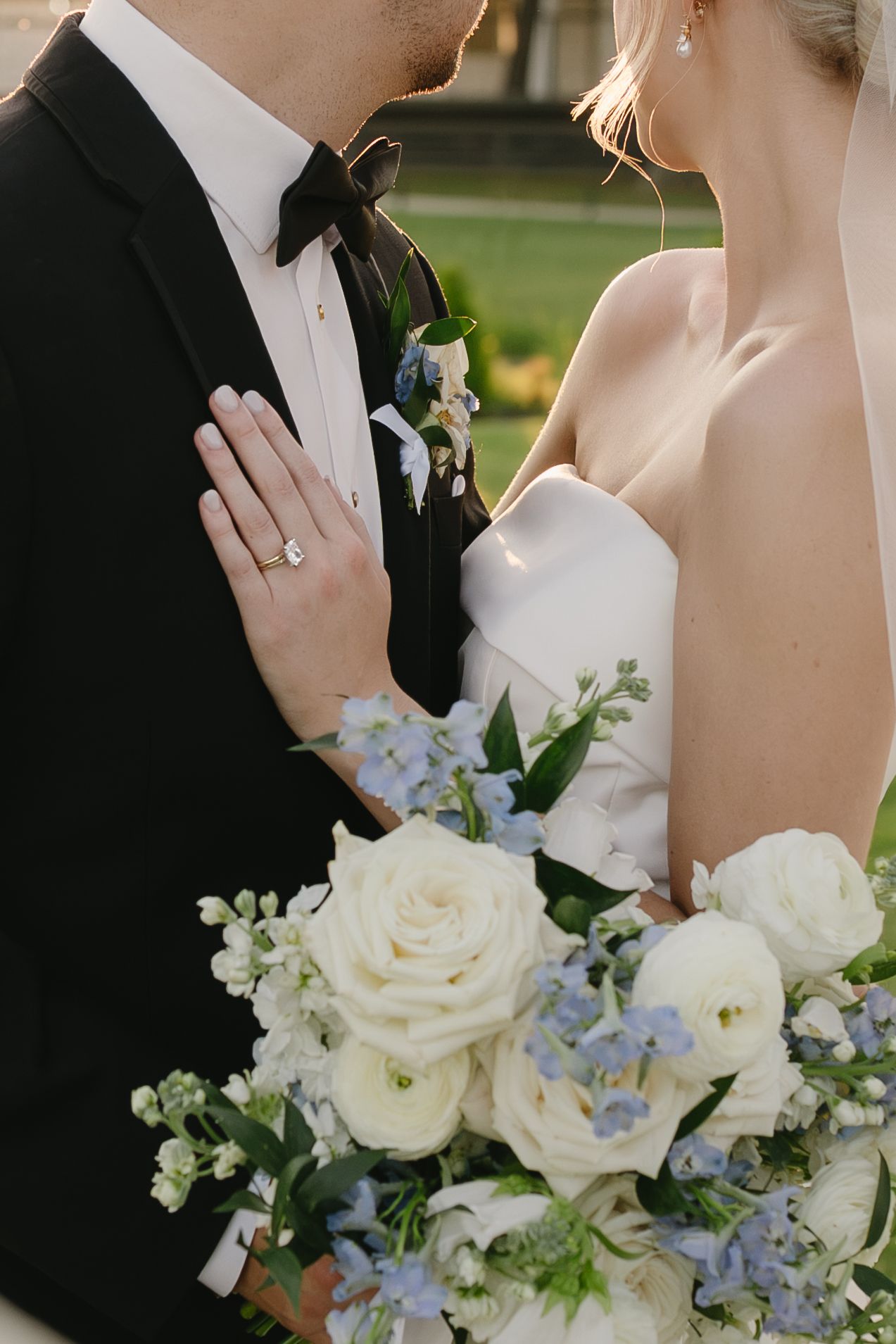 Bride and groom share a romantic moment, her hand resting on his tuxedo, holding a white and blue floral bouquet.