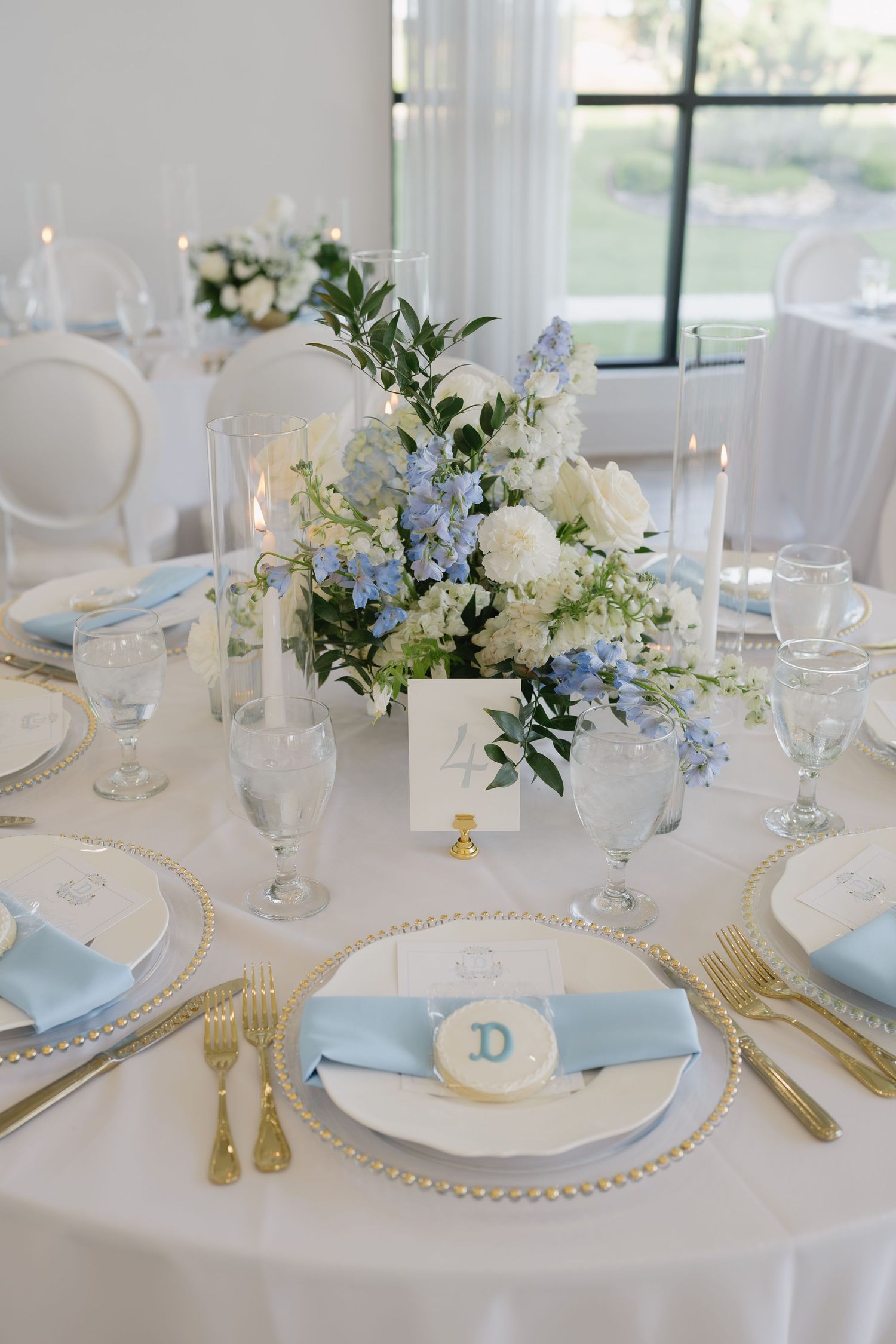 Elegant wedding table setting with blue napkins, gold flatware, floral centerpiece, and personalized place settings.