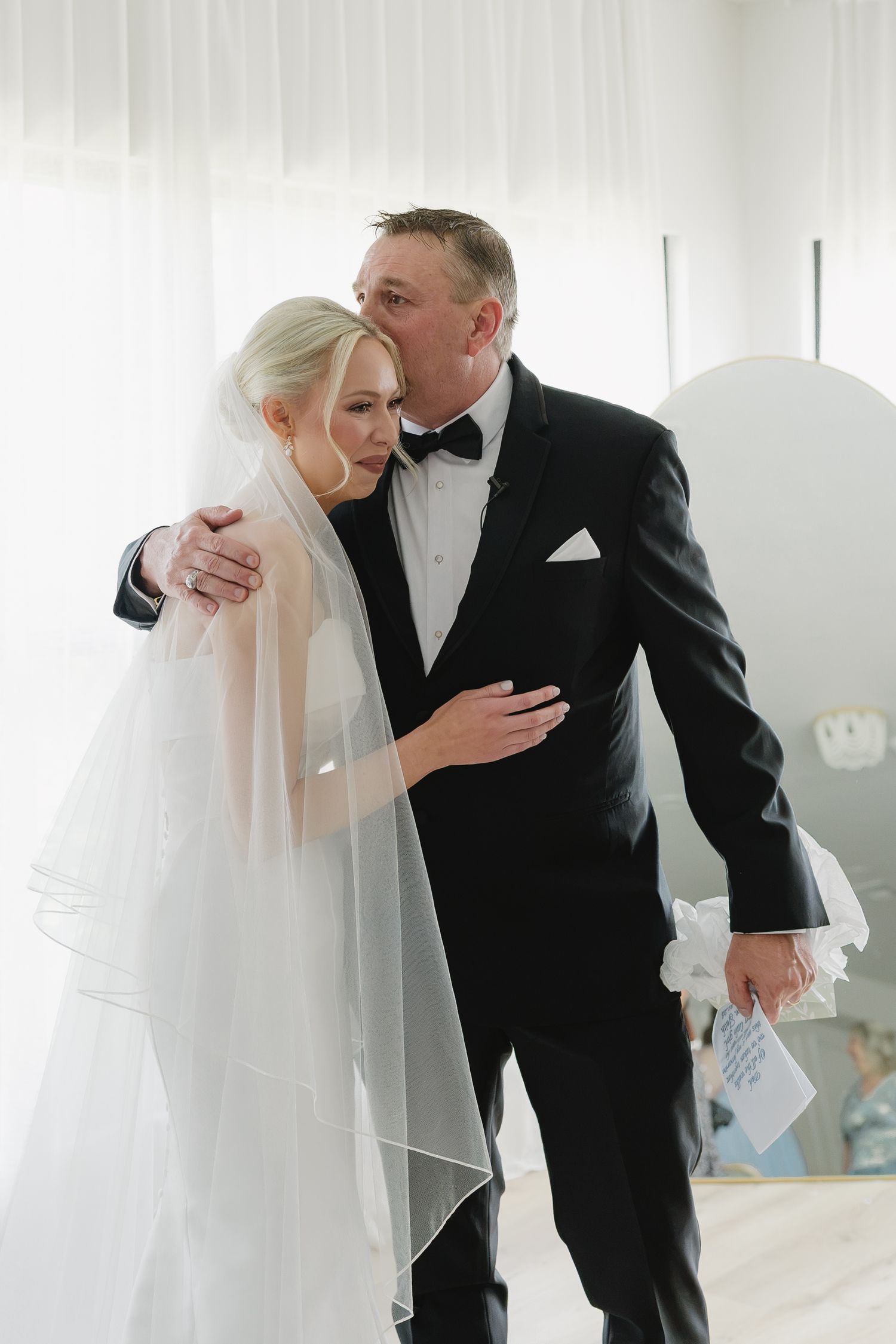 A bride in a white veil embraces a man in a black tuxedo in an emotional wedding day moment.