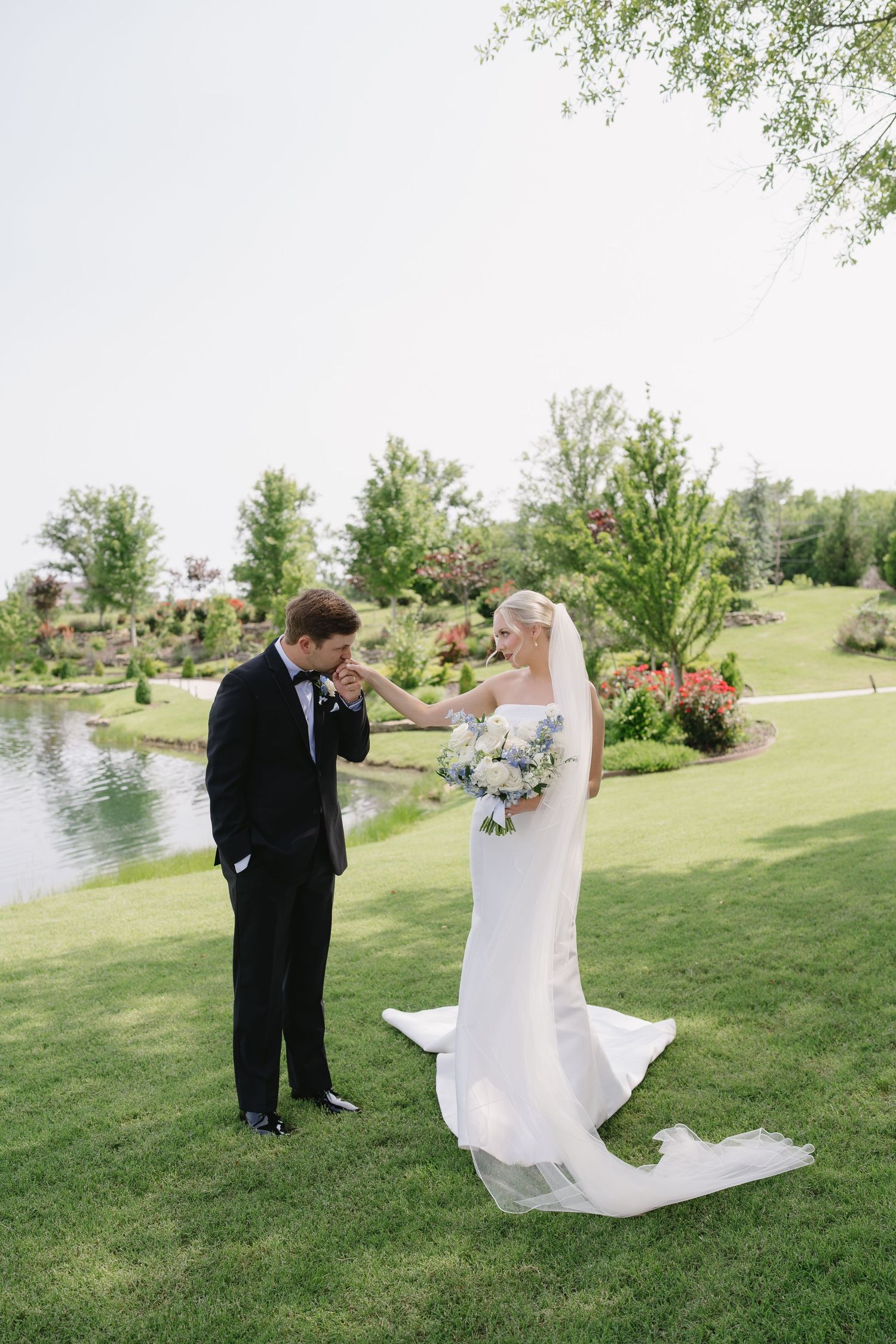 Groom in black tuxedo kisses bride's hand on lush green lawn with pond and garden backdrop.