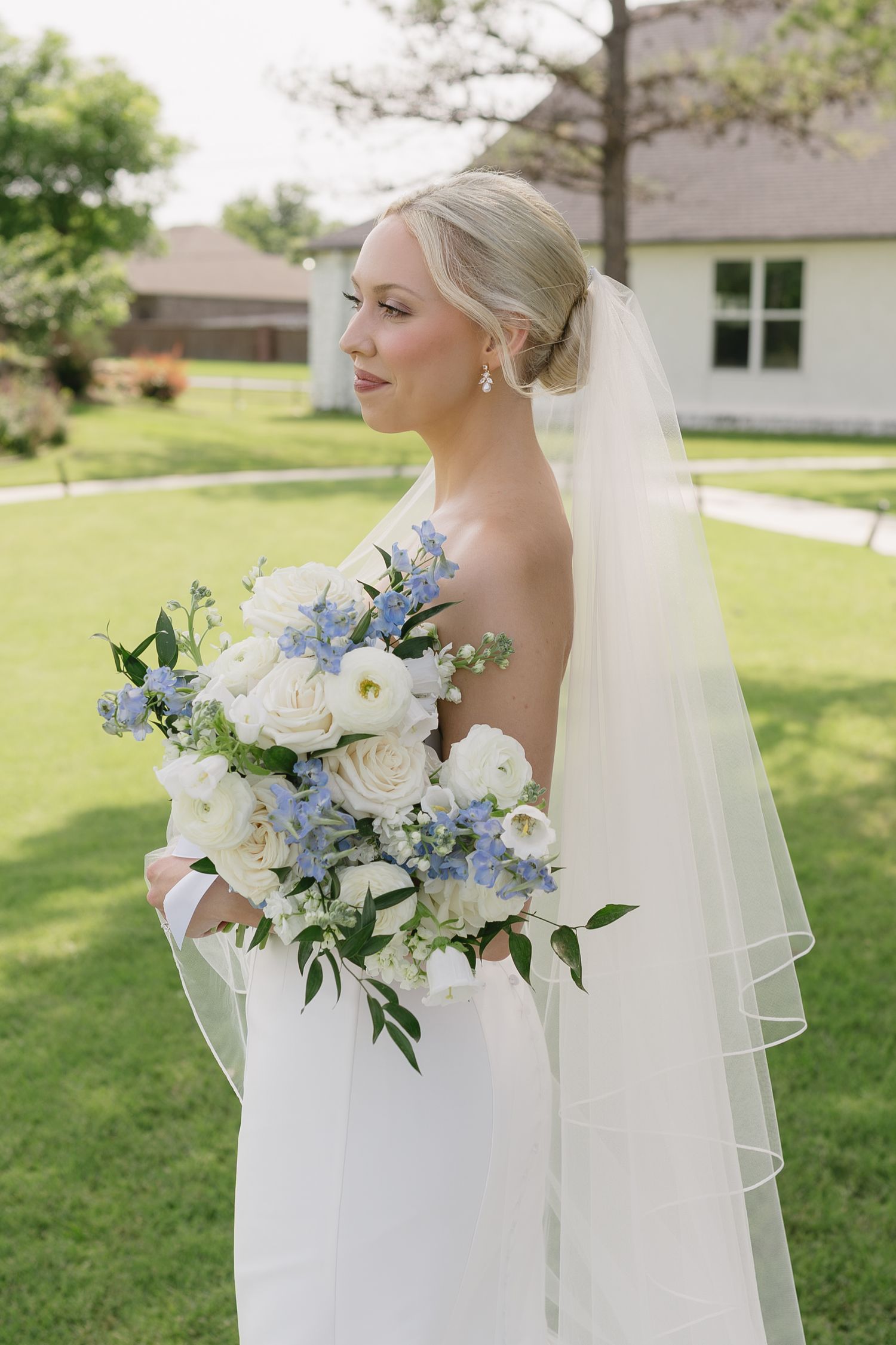 Elegant bride in white strapless gown and veil holding blue and white floral bouquet outdoors.