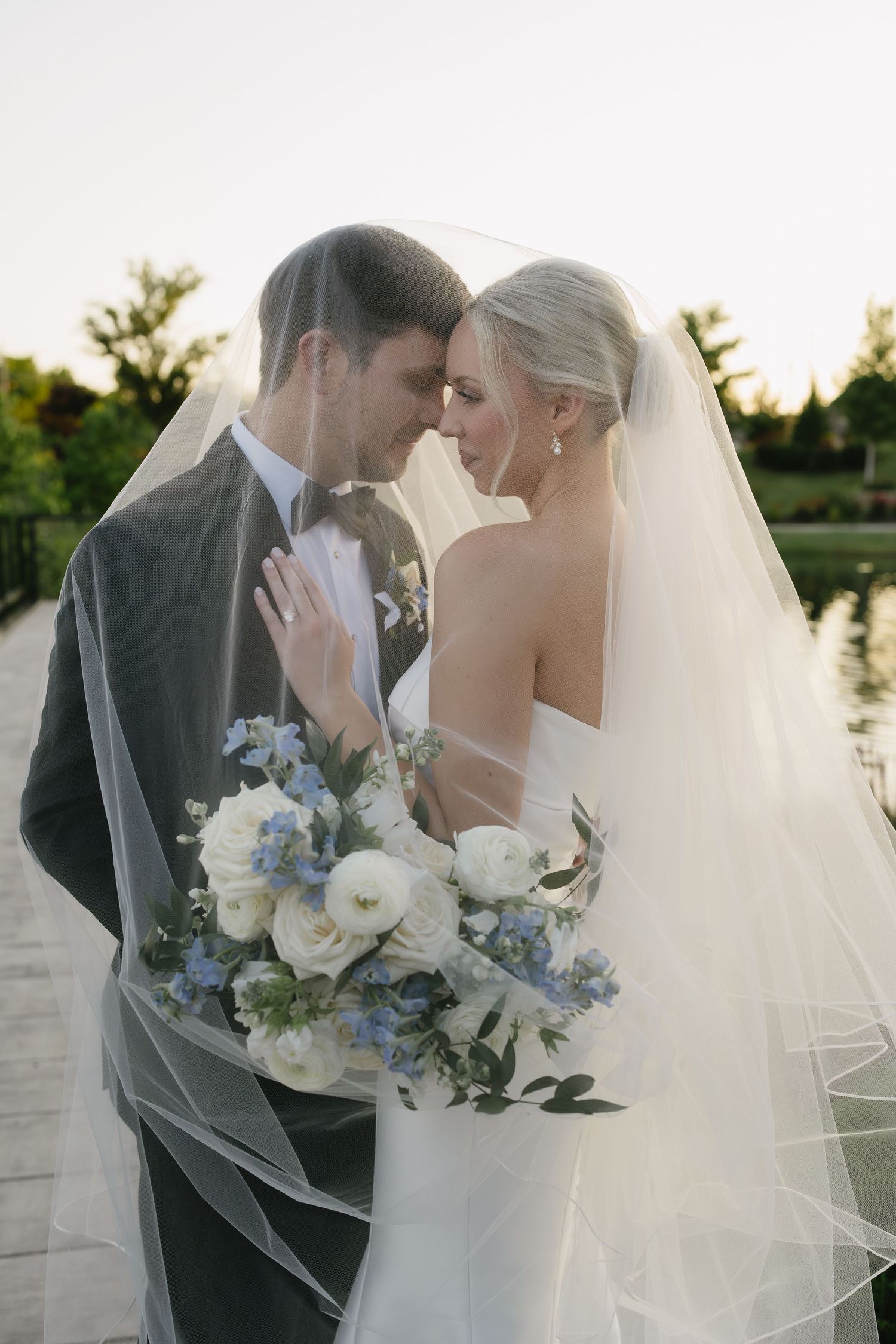 Bride and groom share an intimate moment under a wedding veil, holding a white and blue floral bouquet outdoors.
