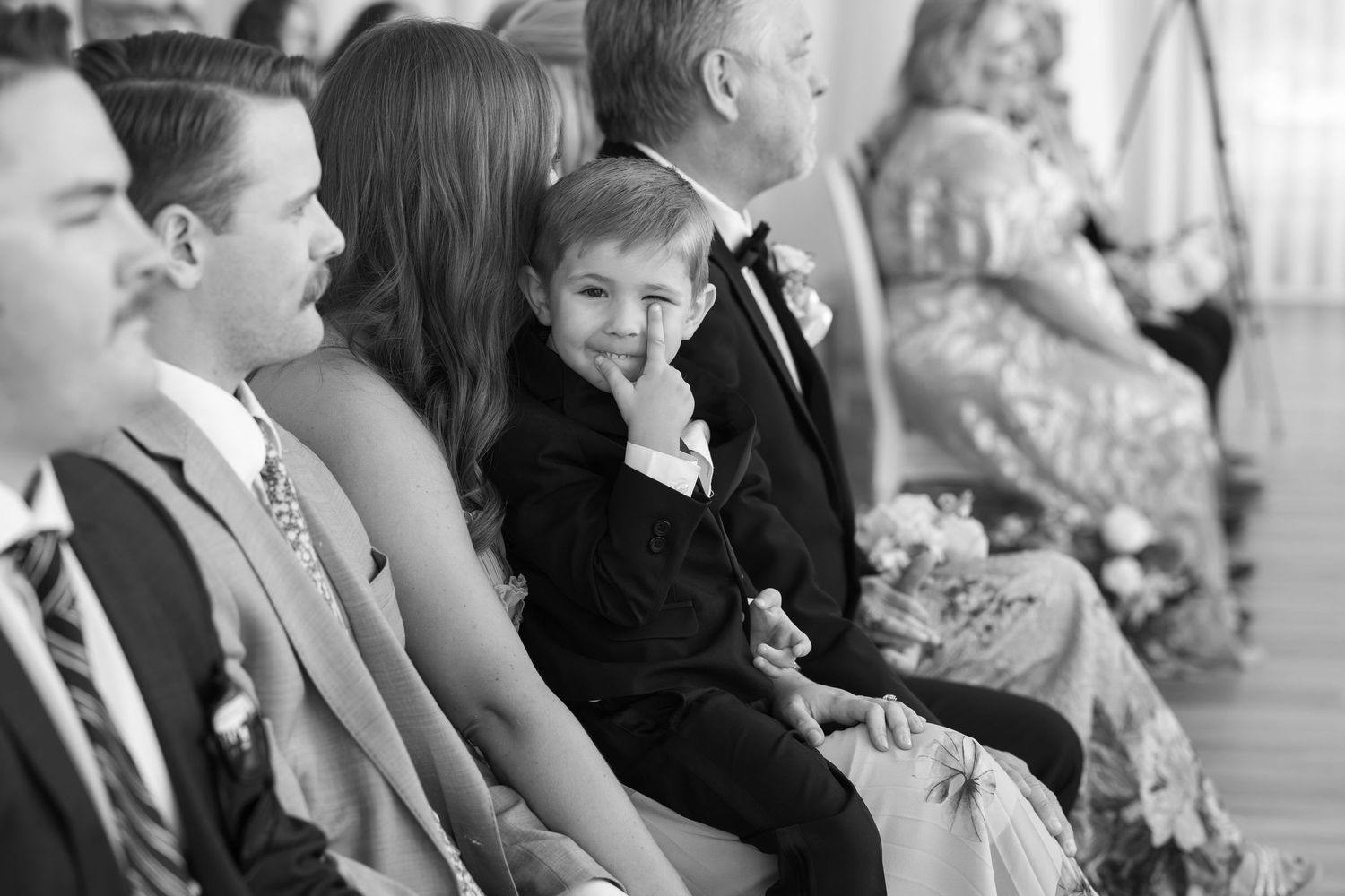 A young boy in a suit smiles at a wedding ceremony, seated among elegantly dressed guests in black and white.