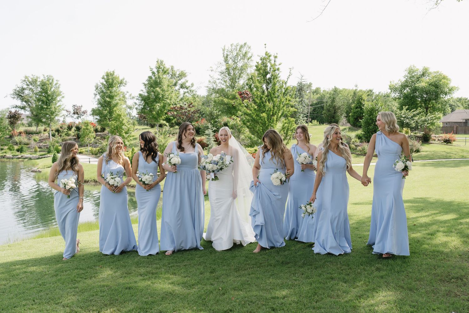 Bride in white gown poses with bridesmaids in light blue dresses outdoors near a pond on a sunny day.