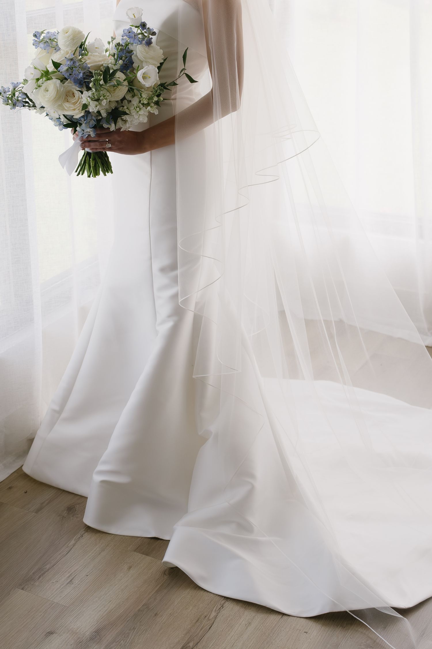 Bride in a white satin mermaid gown with veil holding a blue and white floral bouquet by a bright window.