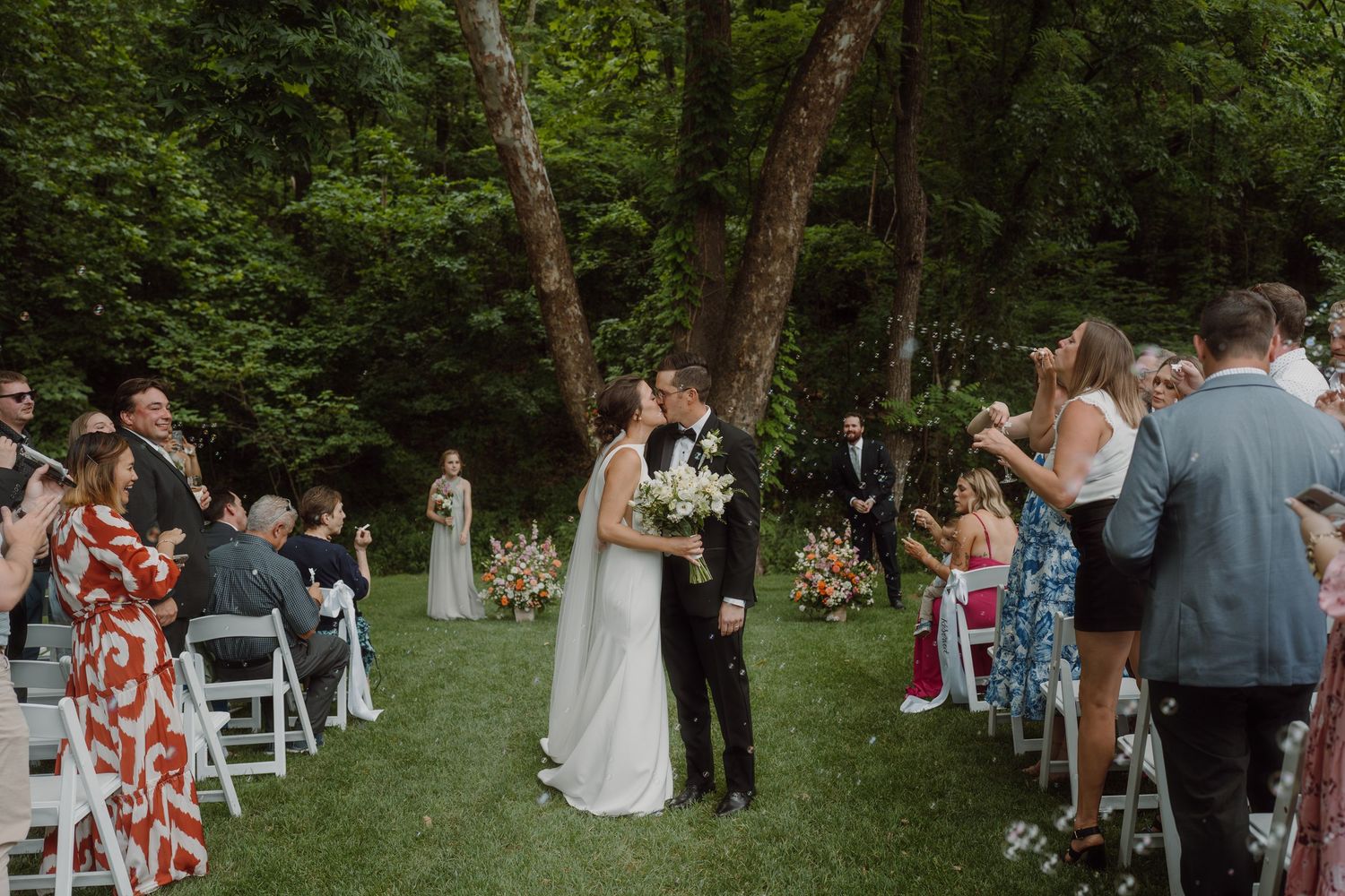 Wedding guests celebrate during an outdoor ceremony in a lush green forest setting.