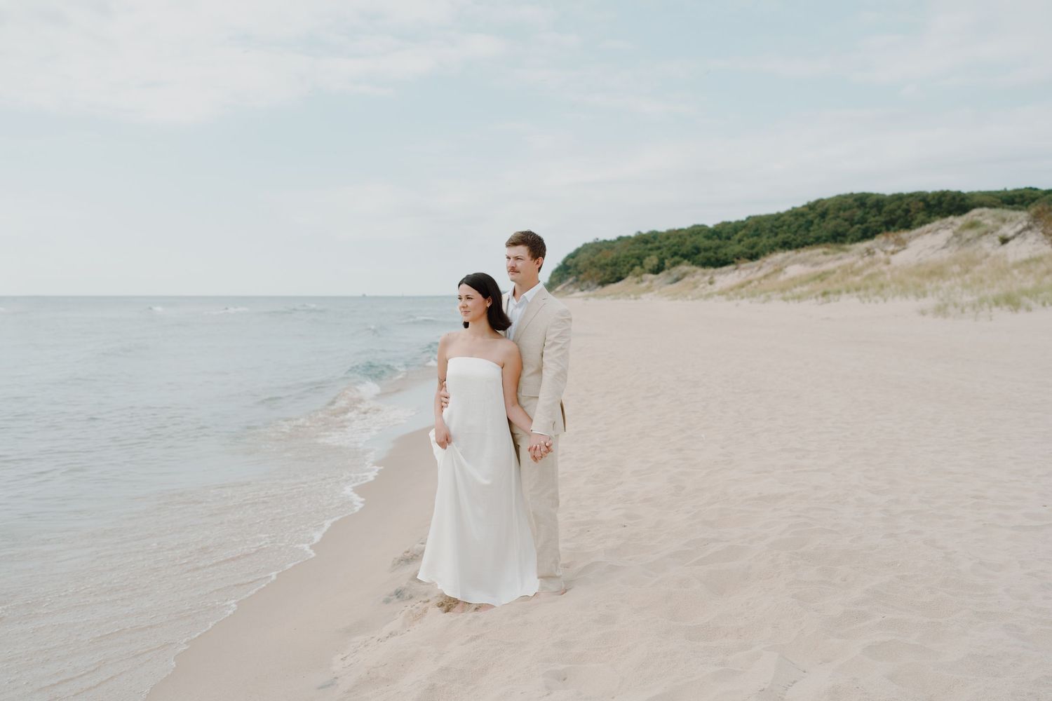 A couple walks hand in hand along a serene beach with gentle waves and sand dunes.