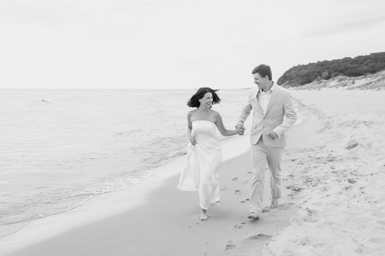 Two people in light colored clothing run playfully along a misty beach shoreline.