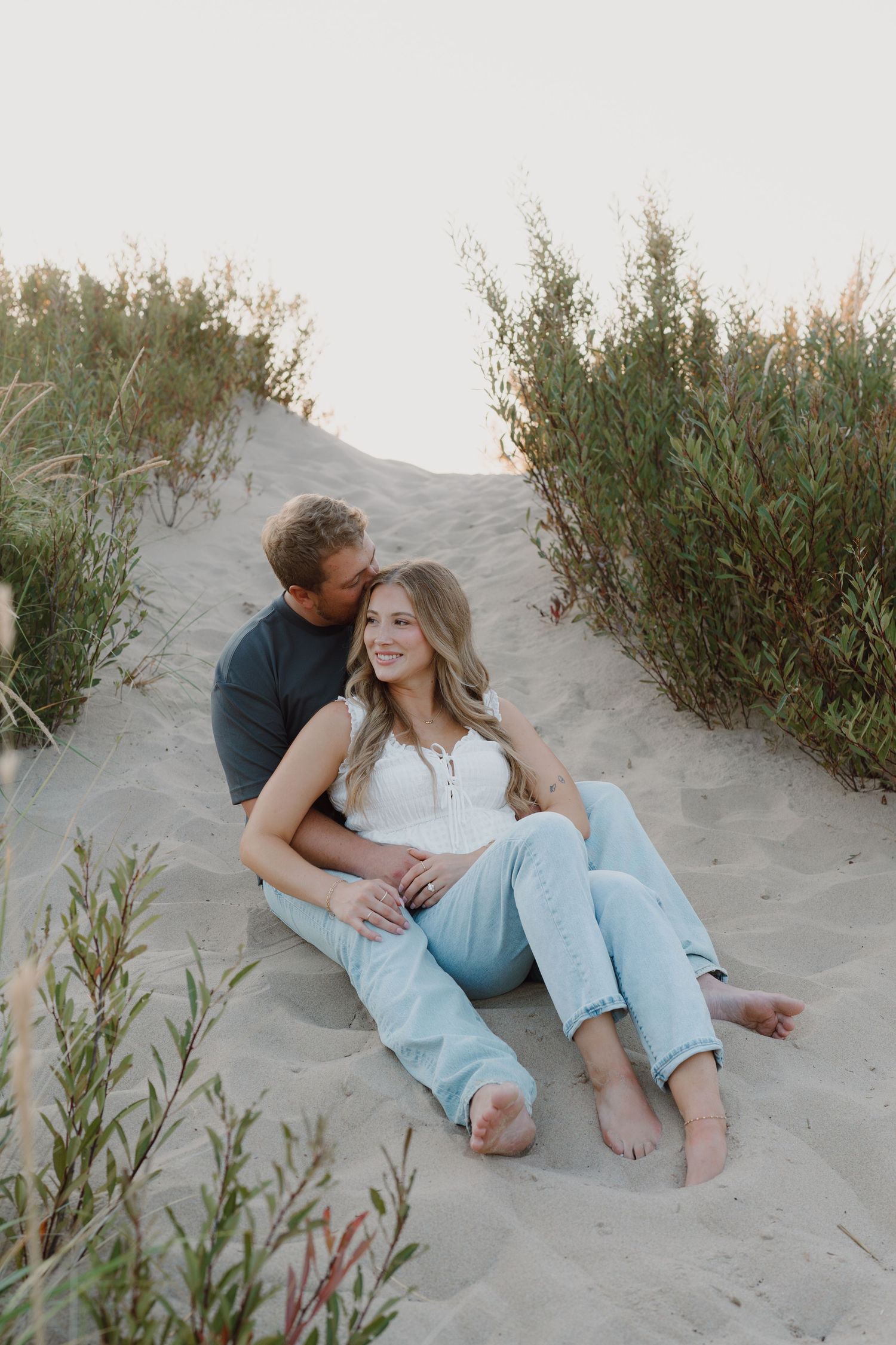 A couple lounges together on sandy dunes during a beach photoshoot at sunset.