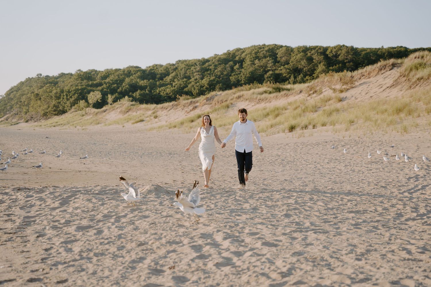 A couple in light clothing takes a romantic beach walk with their small dog.
