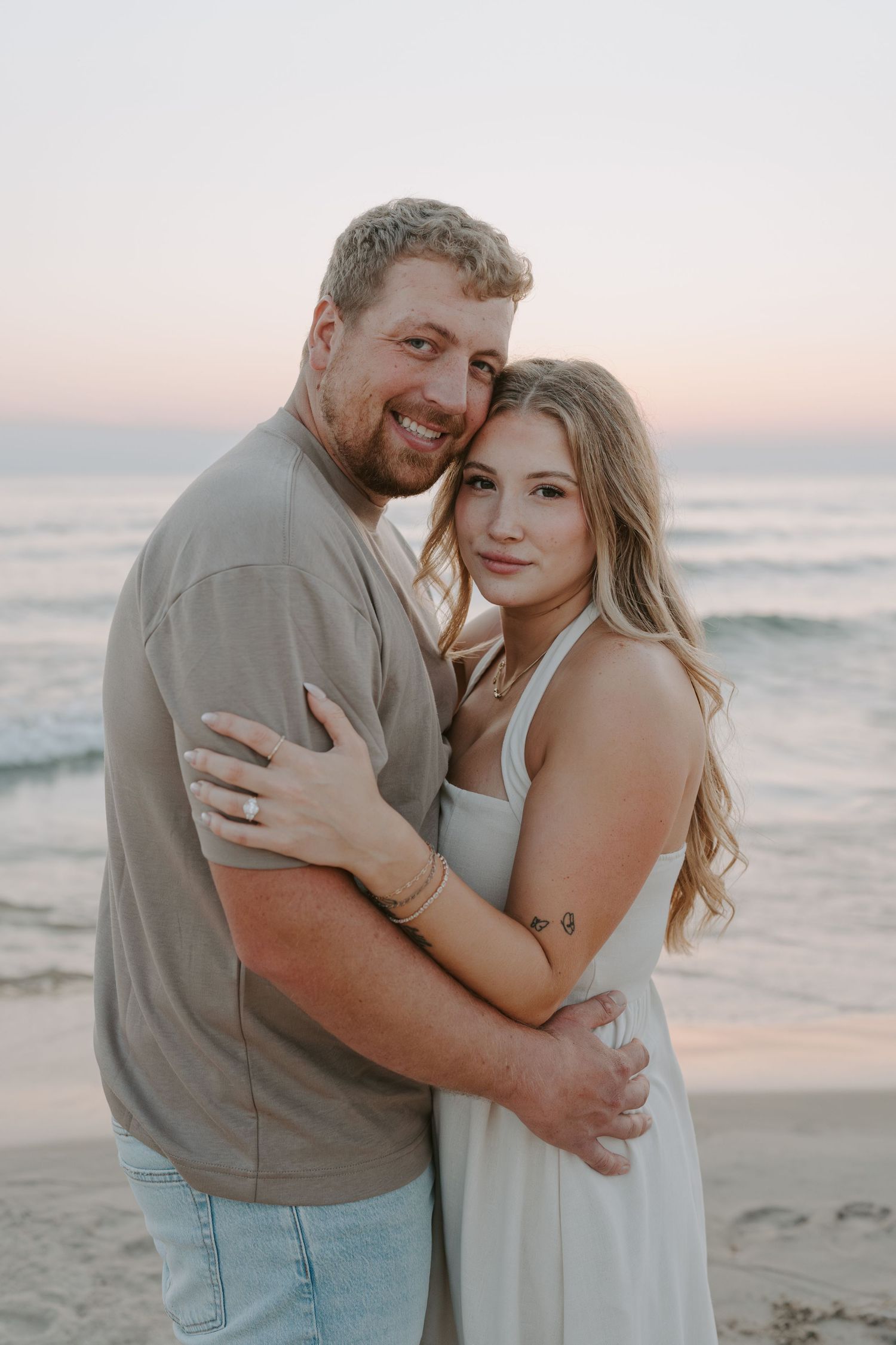 A couple embraces on a beach at sunset wearing casual attire.