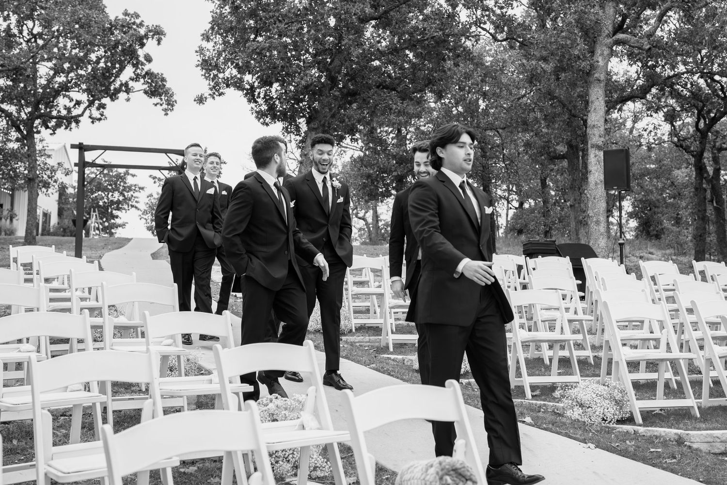 Groomsmen in black suits walk between rows of white chairs at an outdoor wedding ceremony.