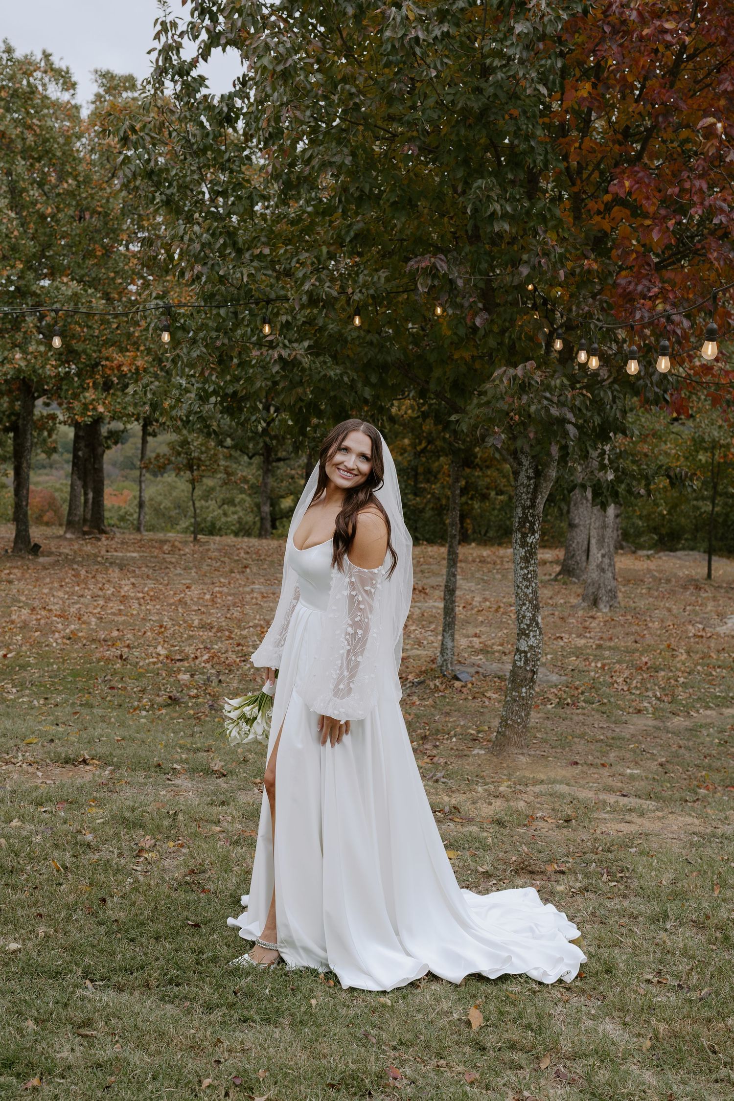 An elegant white wedding gown with lace sleeves photographed in a fall outdoor setting with string lights.