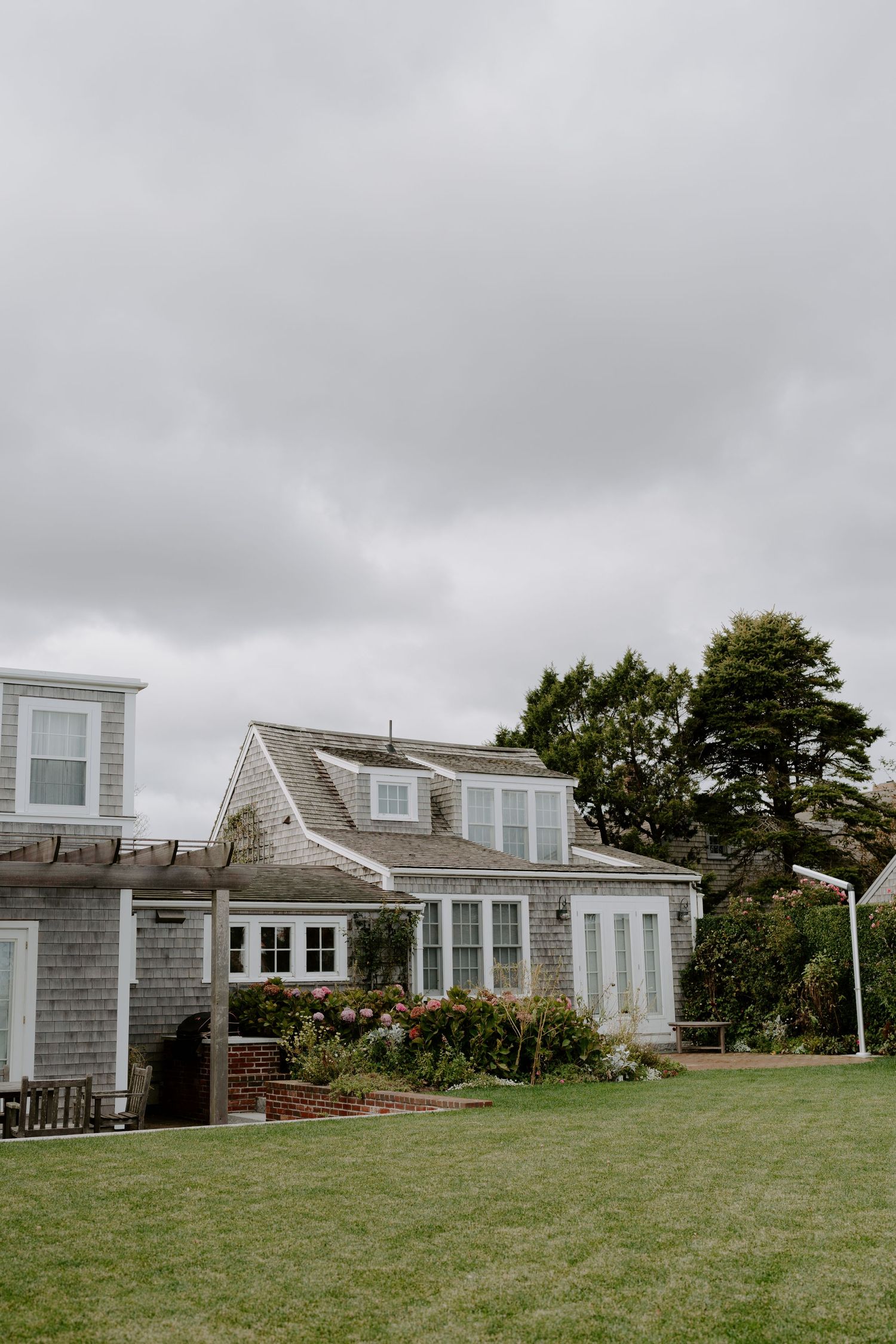 Coastal cottage with gray shingles and white trim features a dormer window and lush green lawn on an overcast day.