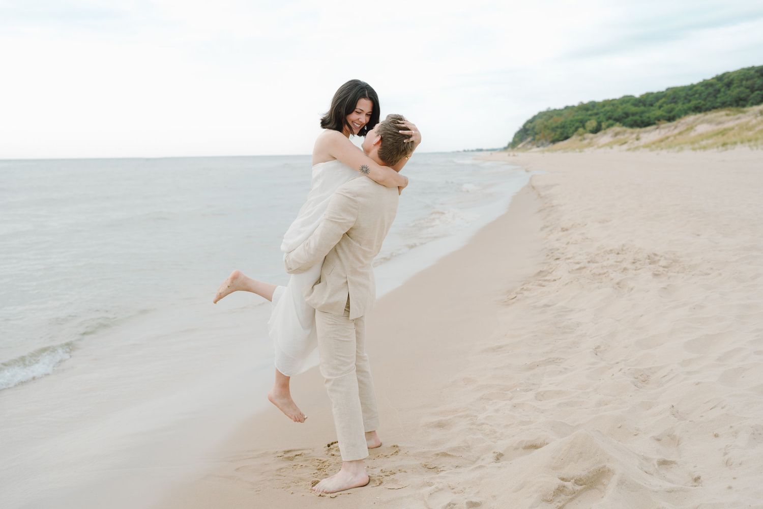 A couple in beige and white outfits joyfully plays on a sandy beach near the water.