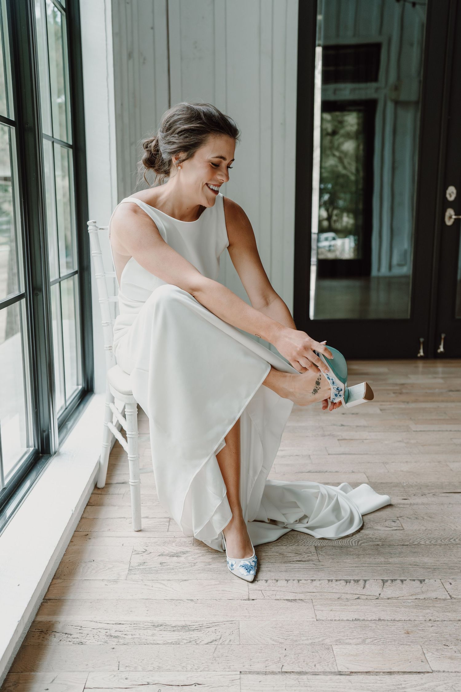 Bride in white dress putting on light blue shoes by large windows.
