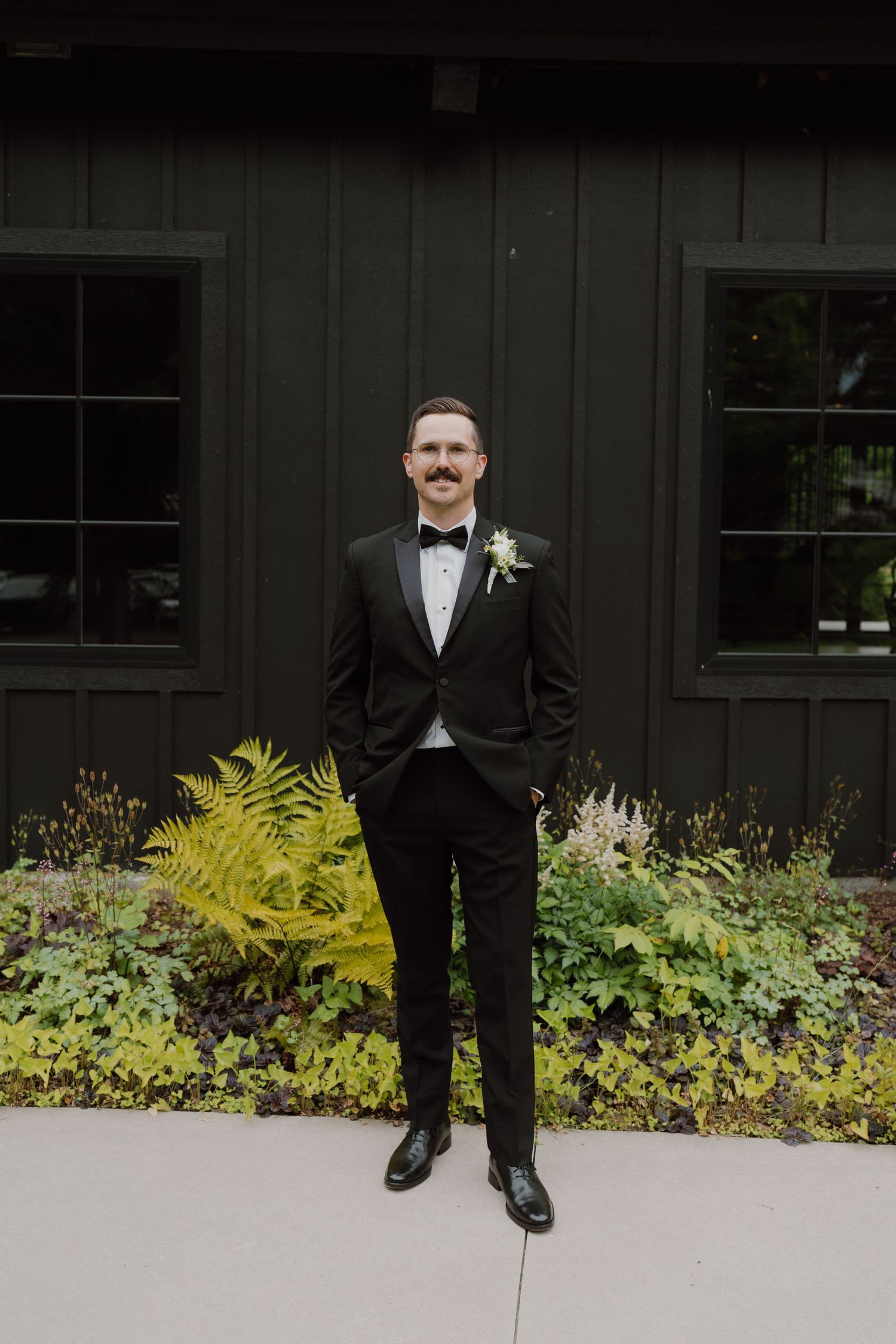 Full length portrait of groom in formal attire against dark wall with garden landscaping.