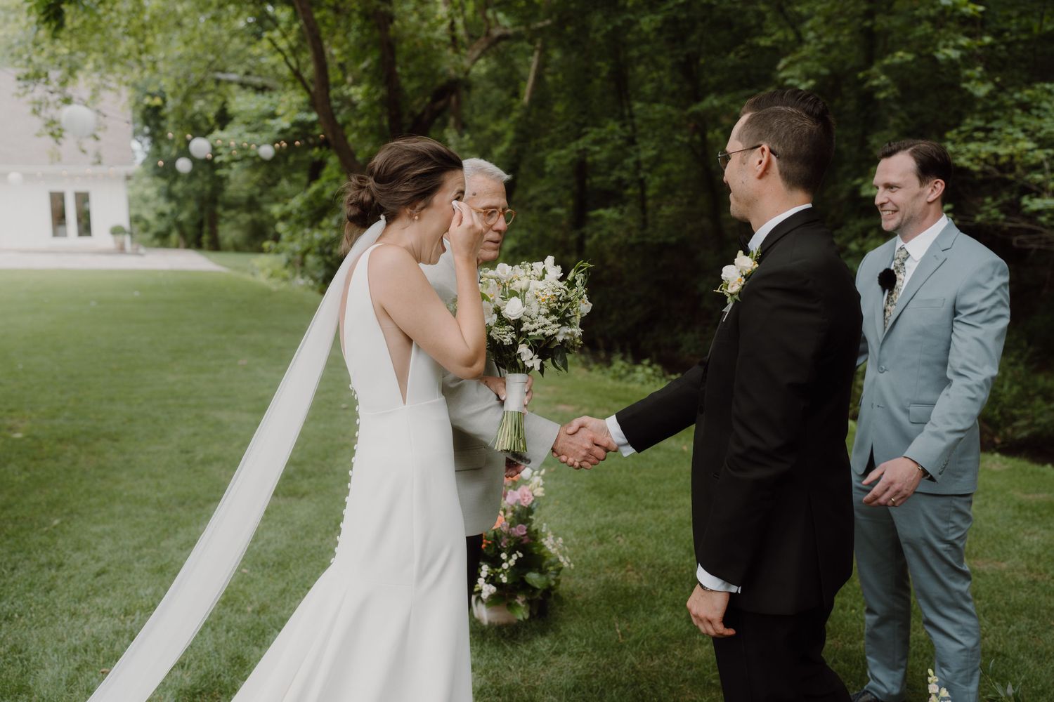 Wedding party members interact during an outdoor ceremony surrounded by greenery and flowers.