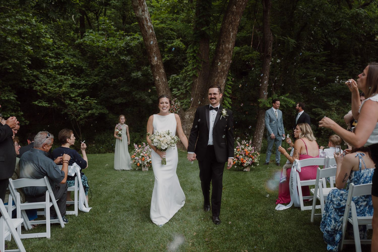 Forest wedding ceremony setup with white chairs and guests arriving for the celebration.