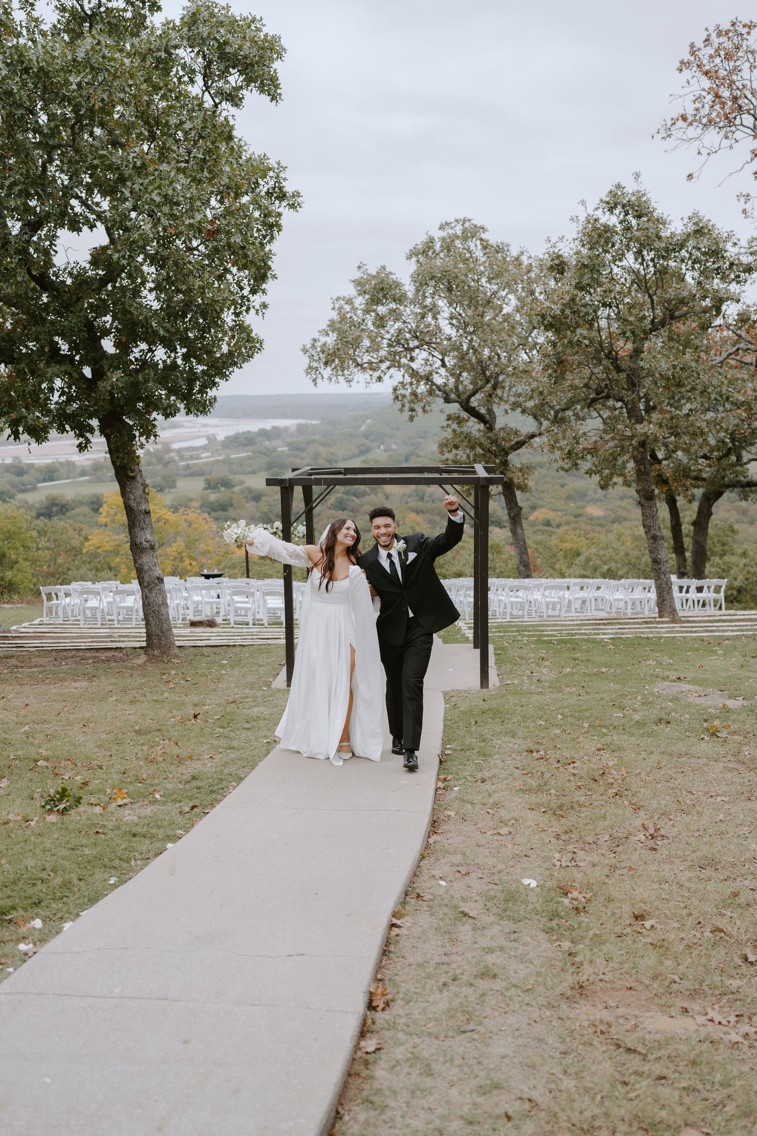 Joyful couple celebrates under wooden arch at outdoor hilltop ceremony site with white chairs.