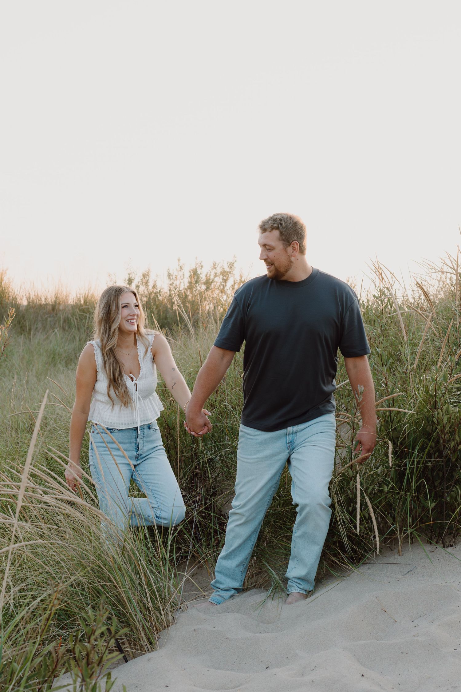 A couple walks together on sandy dunes surrounded by tall beach grass at sunset.