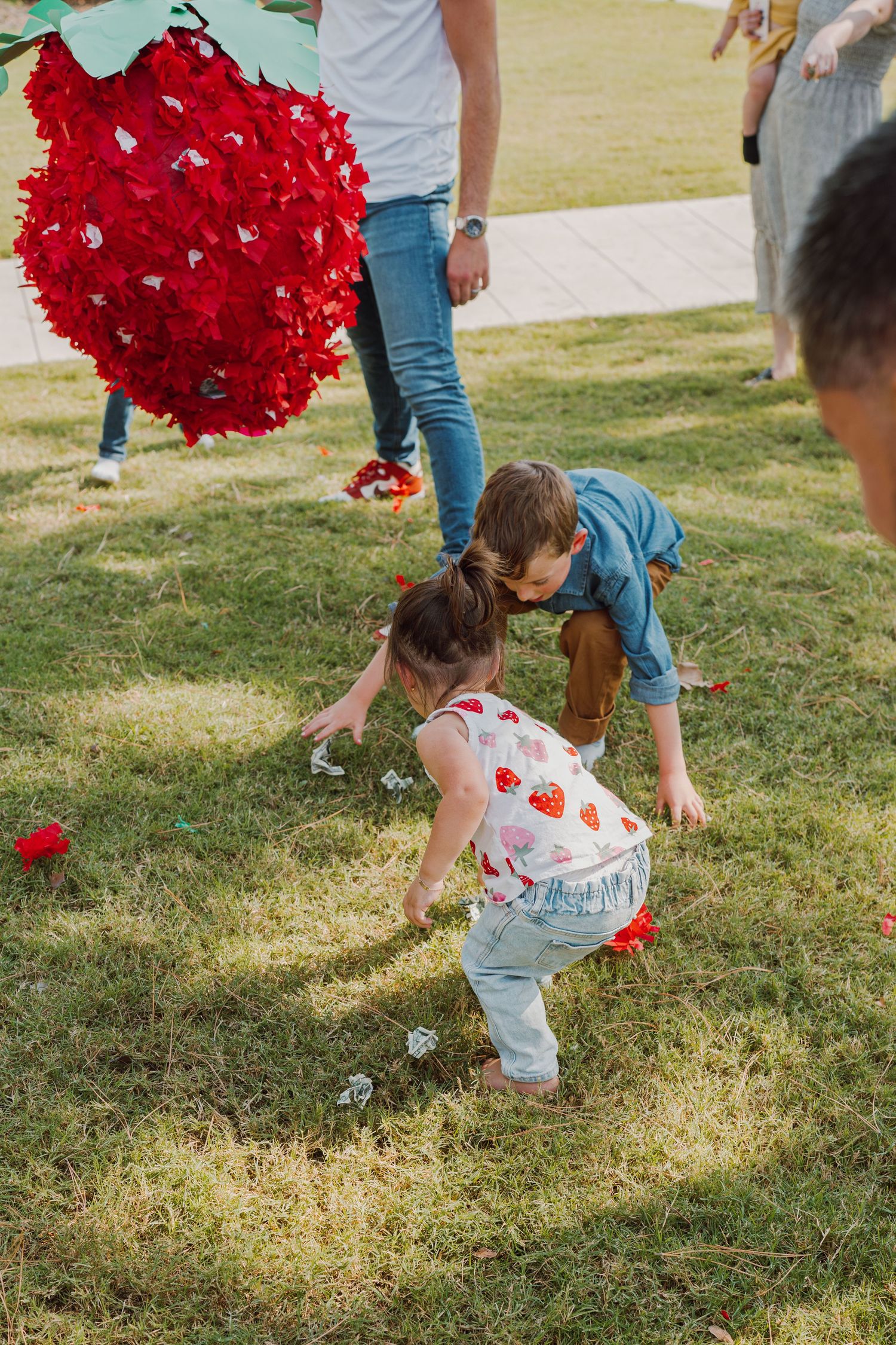 Children picking up scattered red decorative pieces from grass during an outdoor activity.