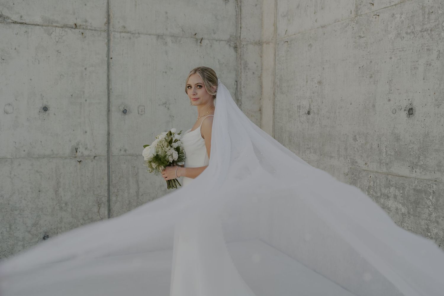Flowing white wedding dress train against industrial concrete wall backdrop.