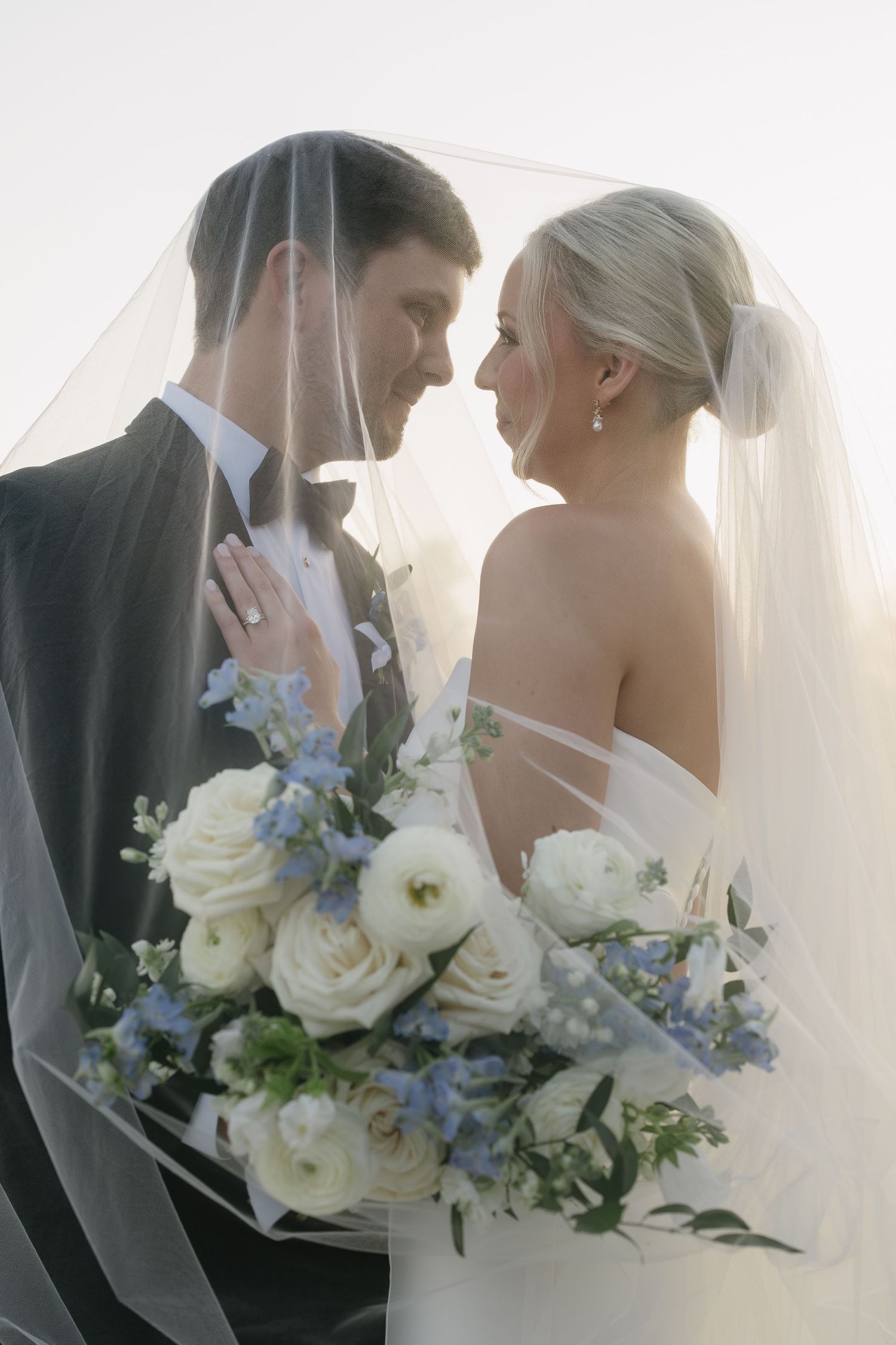 Bride and groom share an intimate moment under a veil, holding a white and blue floral bouquet.