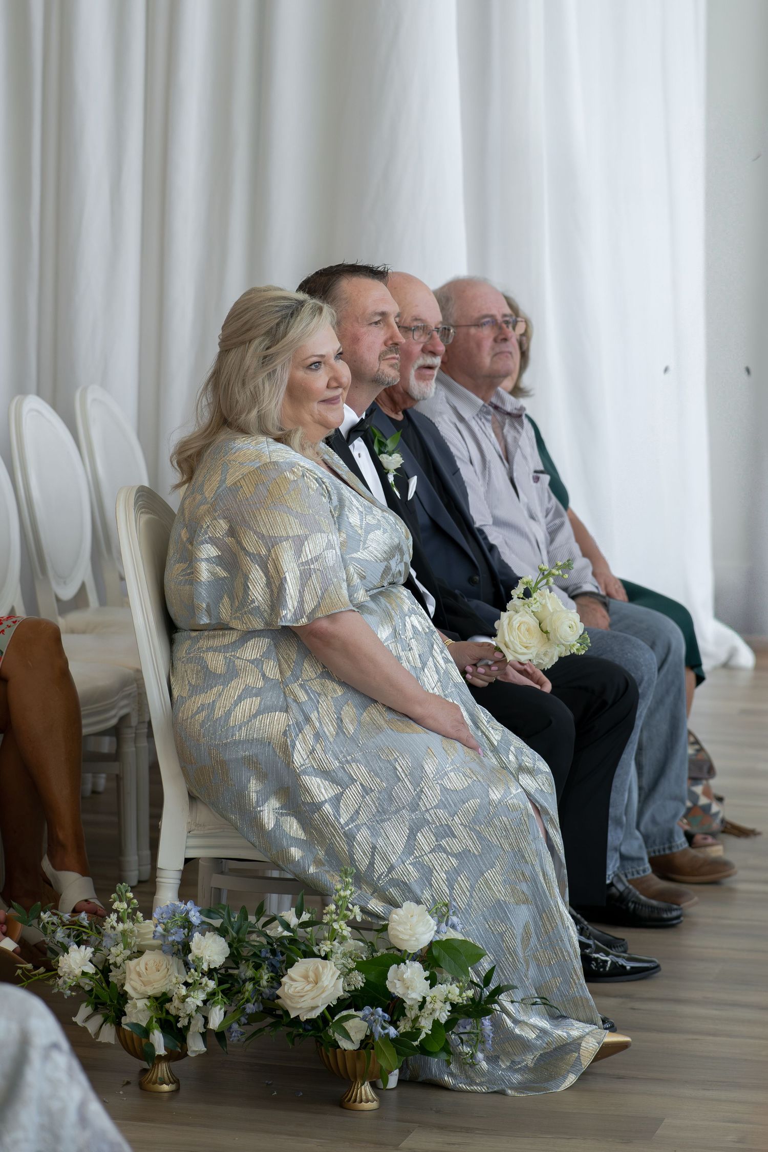 Elegant wedding guests seated in white chairs, woman in silver floral gown holding white roses bouquet at indoor ceremony.