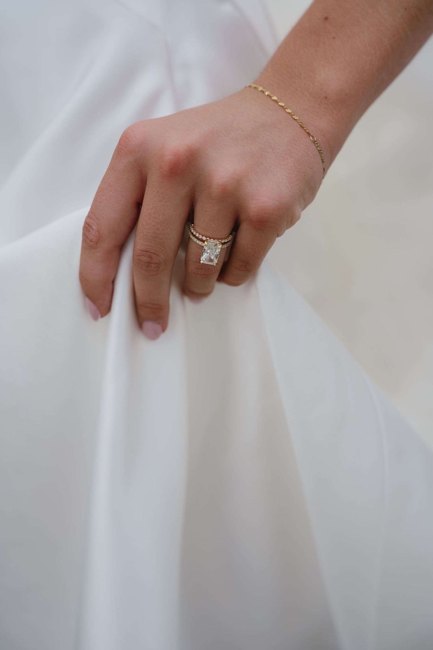 Close-up of a bride's hand wearing a large diamond engagement ring and gold wedding band, holding a white dress.