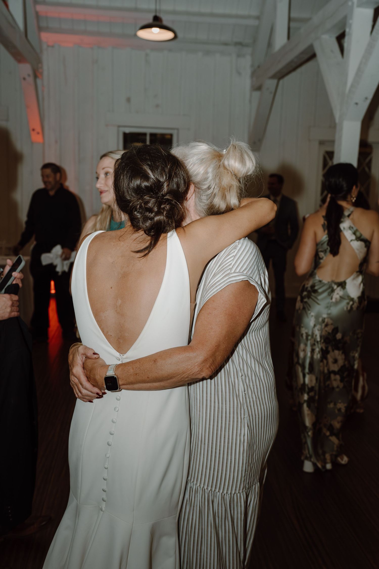 Wedding party members share emotional embraces during a reception in a white-walled venue.