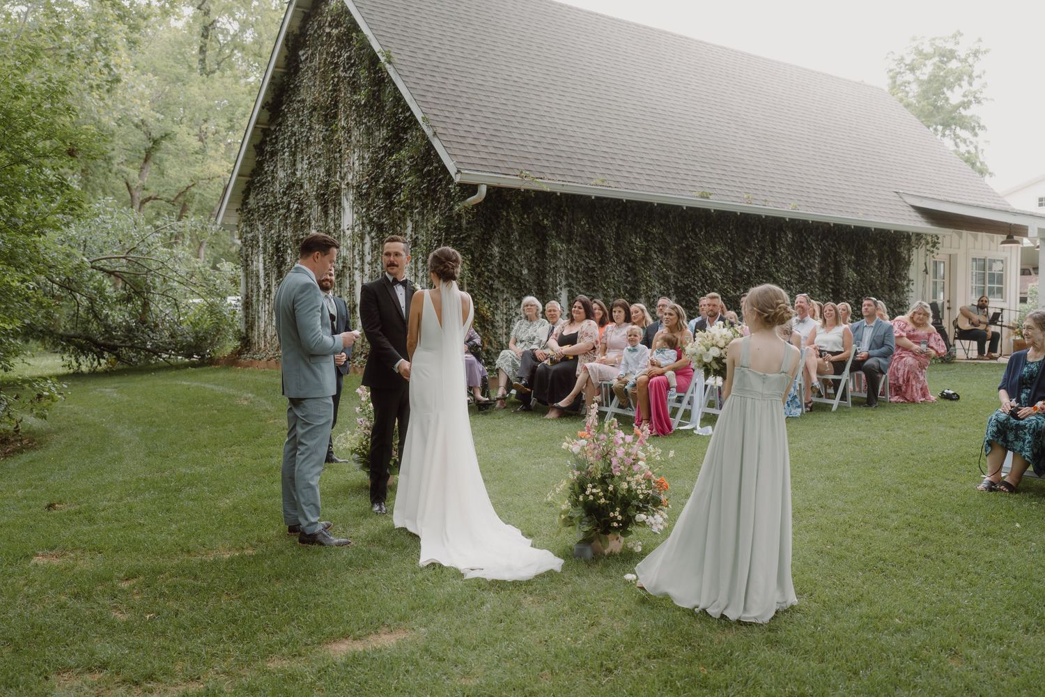 Outdoor wedding ceremony with guests seated on white chairs beneath towering trees.