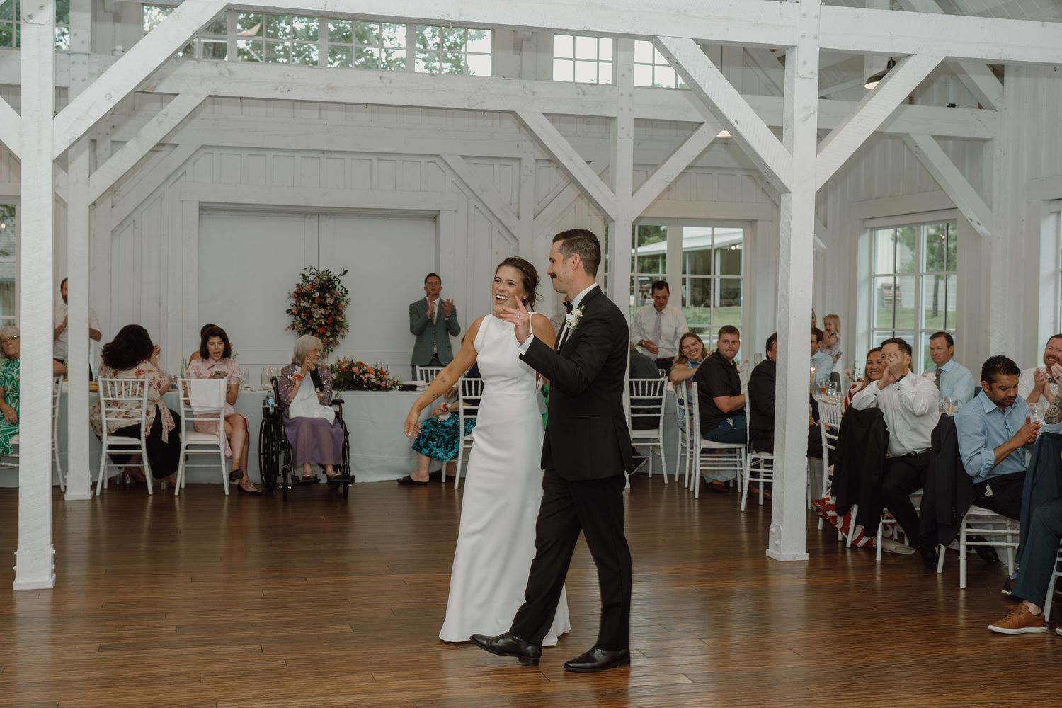 Elegant white barn wedding venue with couples dancing and guests seated at round tables.