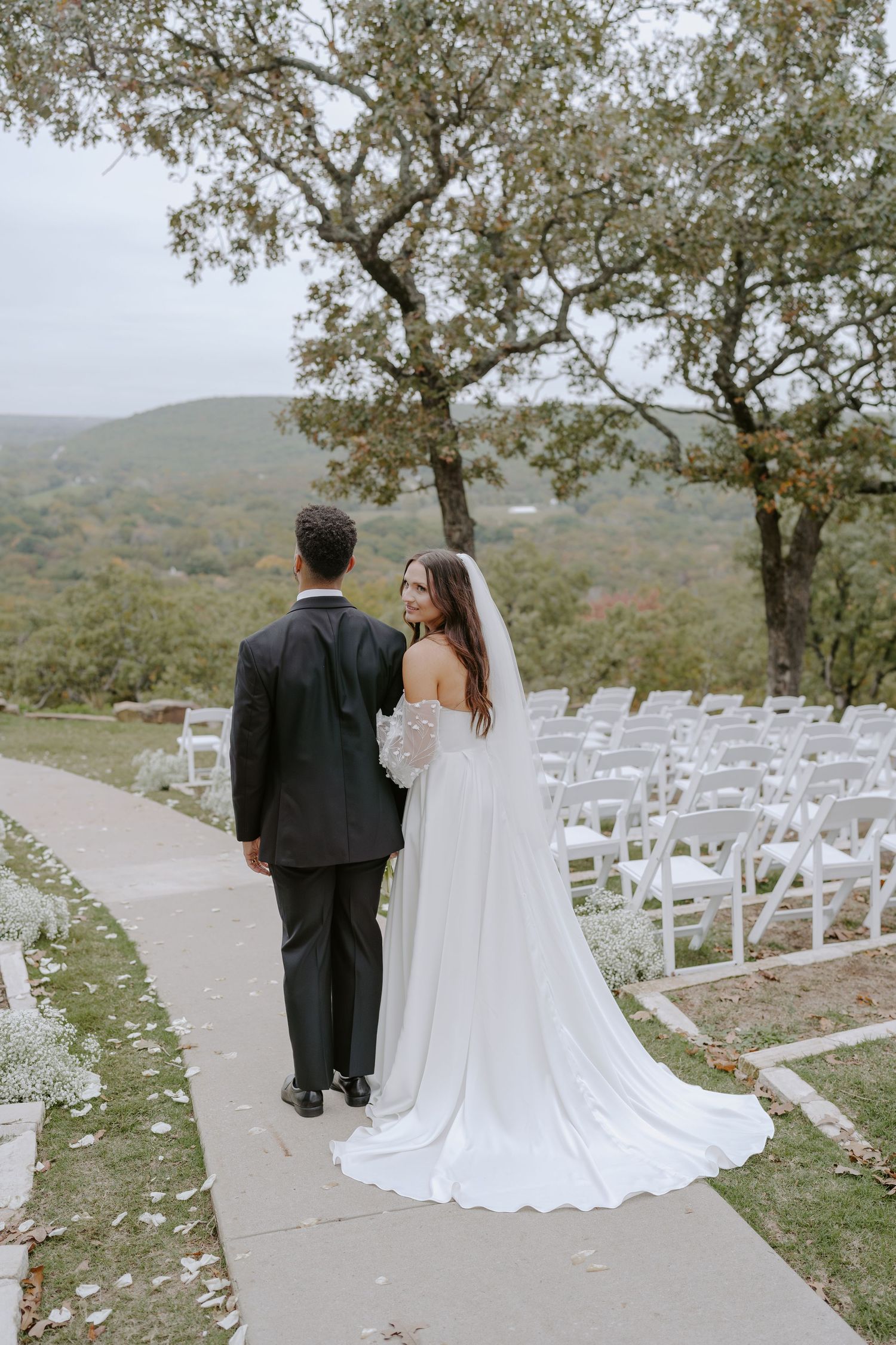 A couple walks along a stone path at their hilltop wedding ceremony site surrounded by autumn trees.